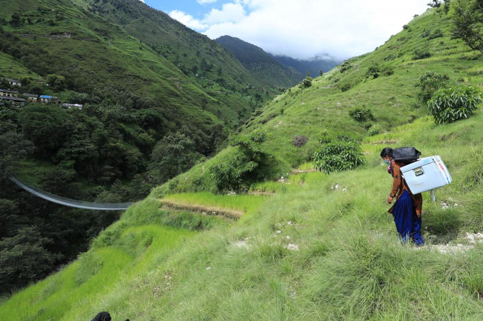 An image of a woman walking with a large box on her back through a gorgeous mountainous region. 