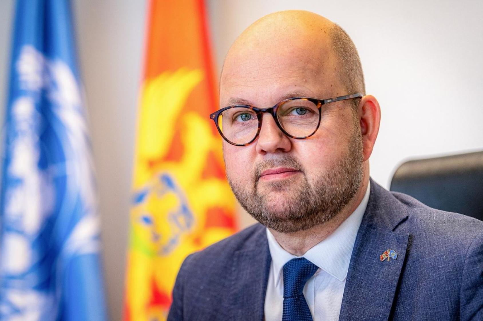 A man in glasses sits in front of two flags while looking directly at the camera.