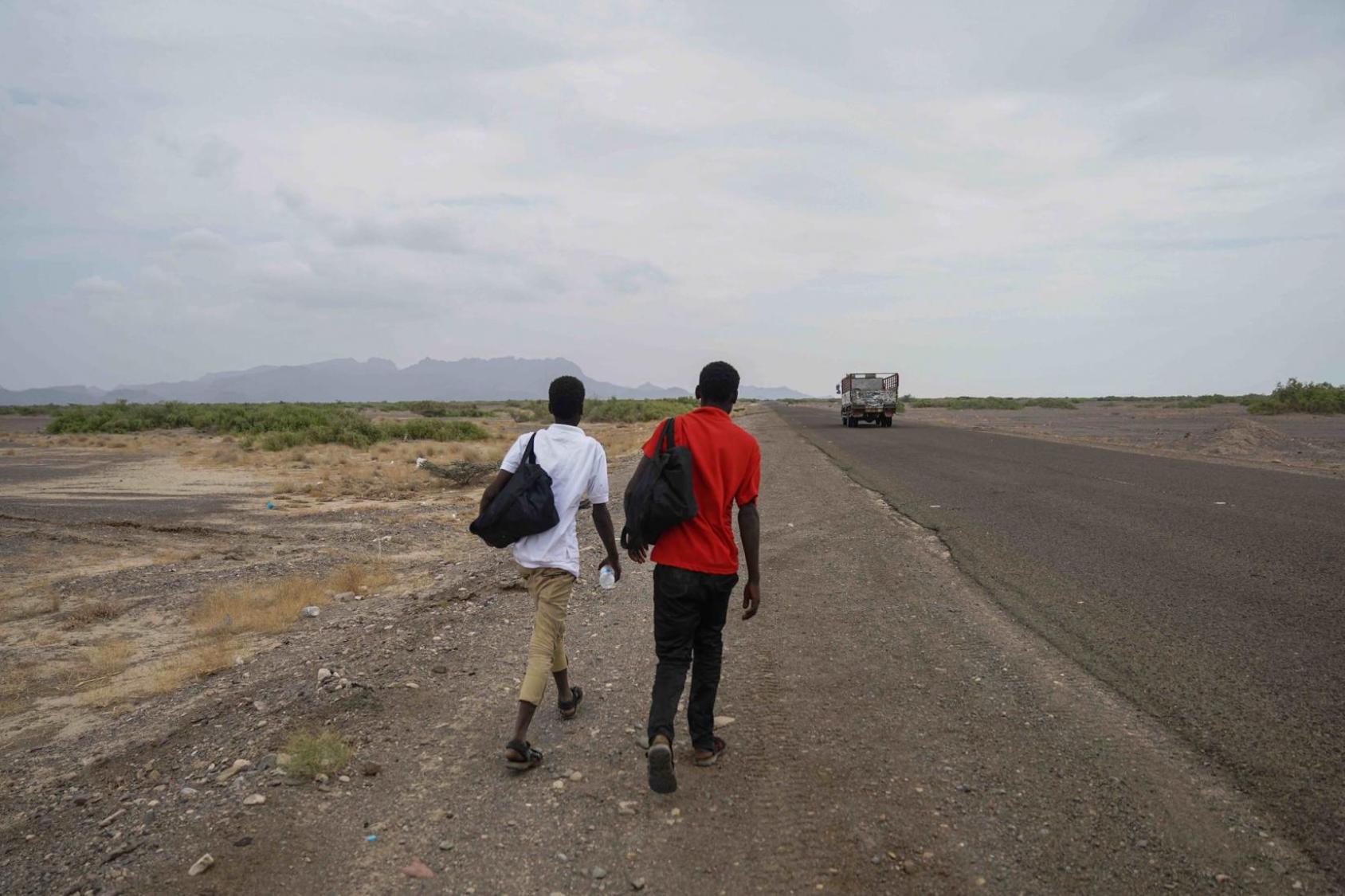 Two people, one in a white shirt and the other in a red shirt, walk on the side of the road on a cloudy day. 