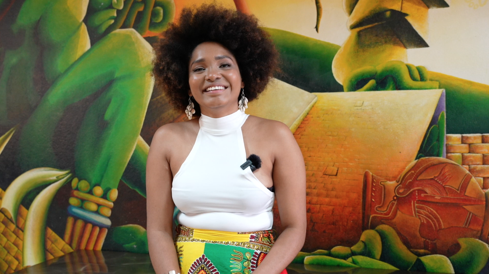 A woman gleefully smiles as she sits in front of a brightly coloured mural. 