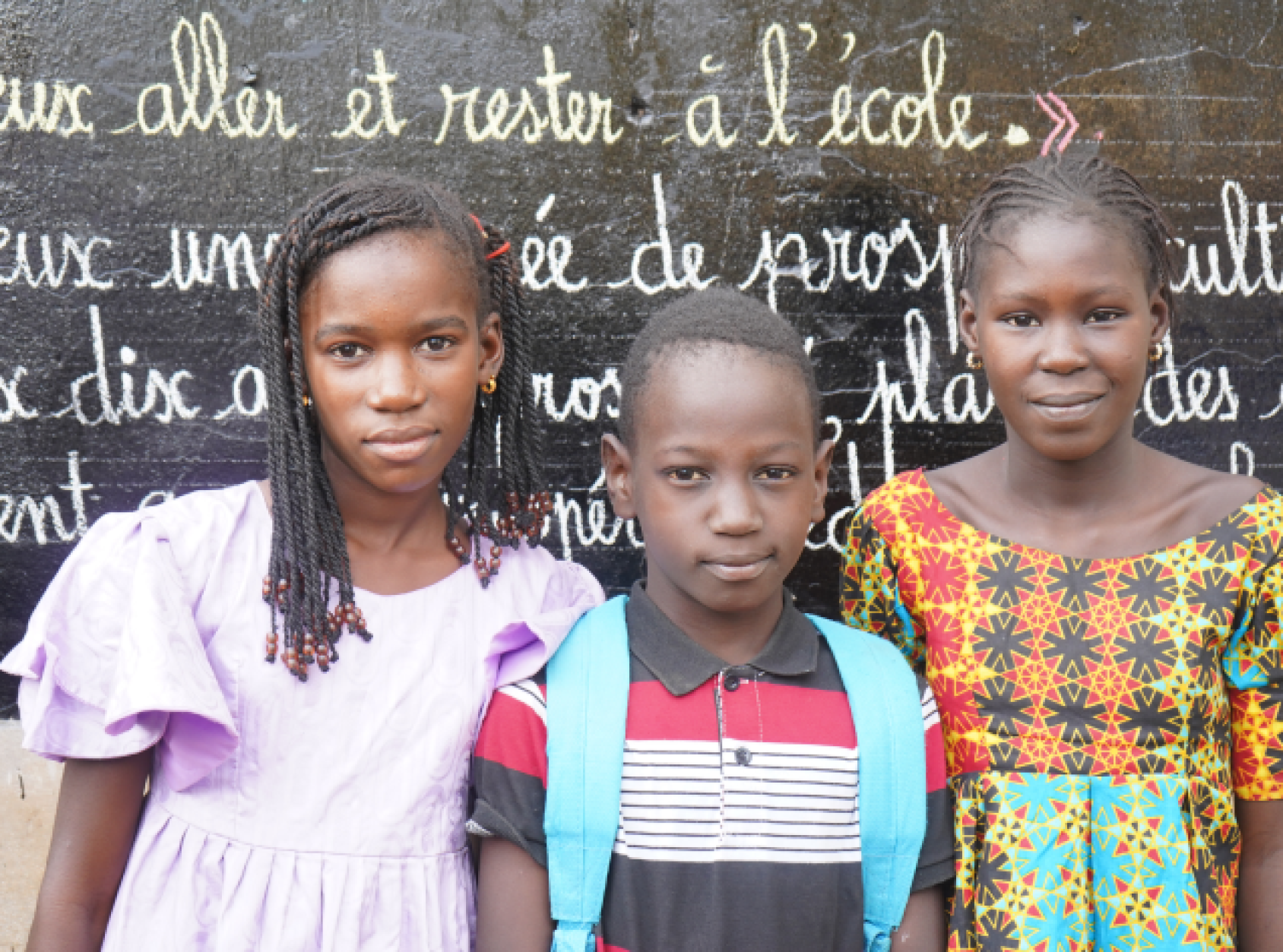Au Sénégal, deux jeunes filles et un garçon se tiennent debout, côte à côte, devant un tableau noir où sont écrites à la craie plusieurs phrases en français, et regardent la caméra en souriant discrètement.