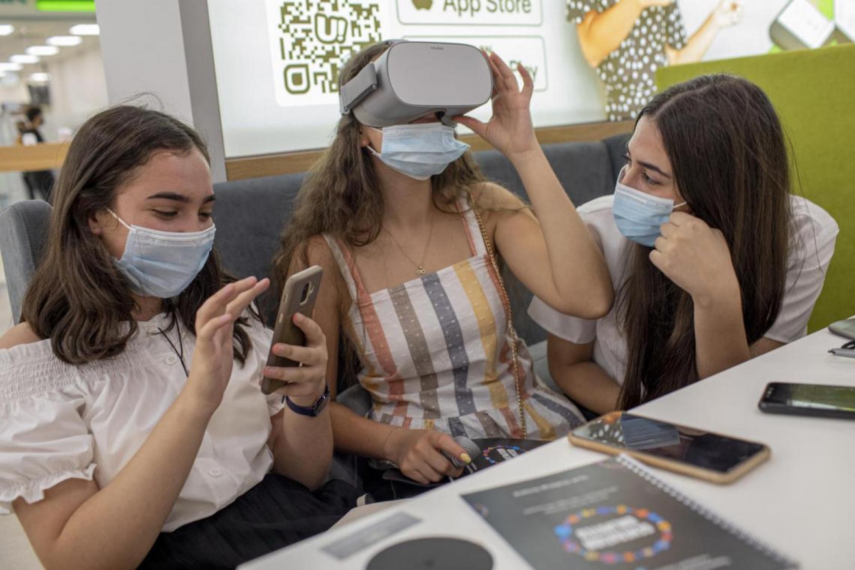 Trois lycéennes arméniennes portant chacune un masque de protection respiratoire sont assises à une table et testent un appareil utilisant la réalité virtuelle. 
