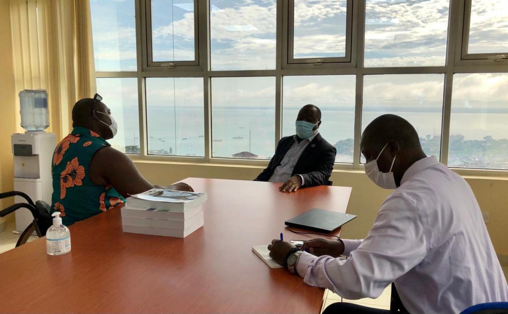 Tres hombres se sientan alrededor de una mesa con mascarillas cerca de una pared con ventanales.