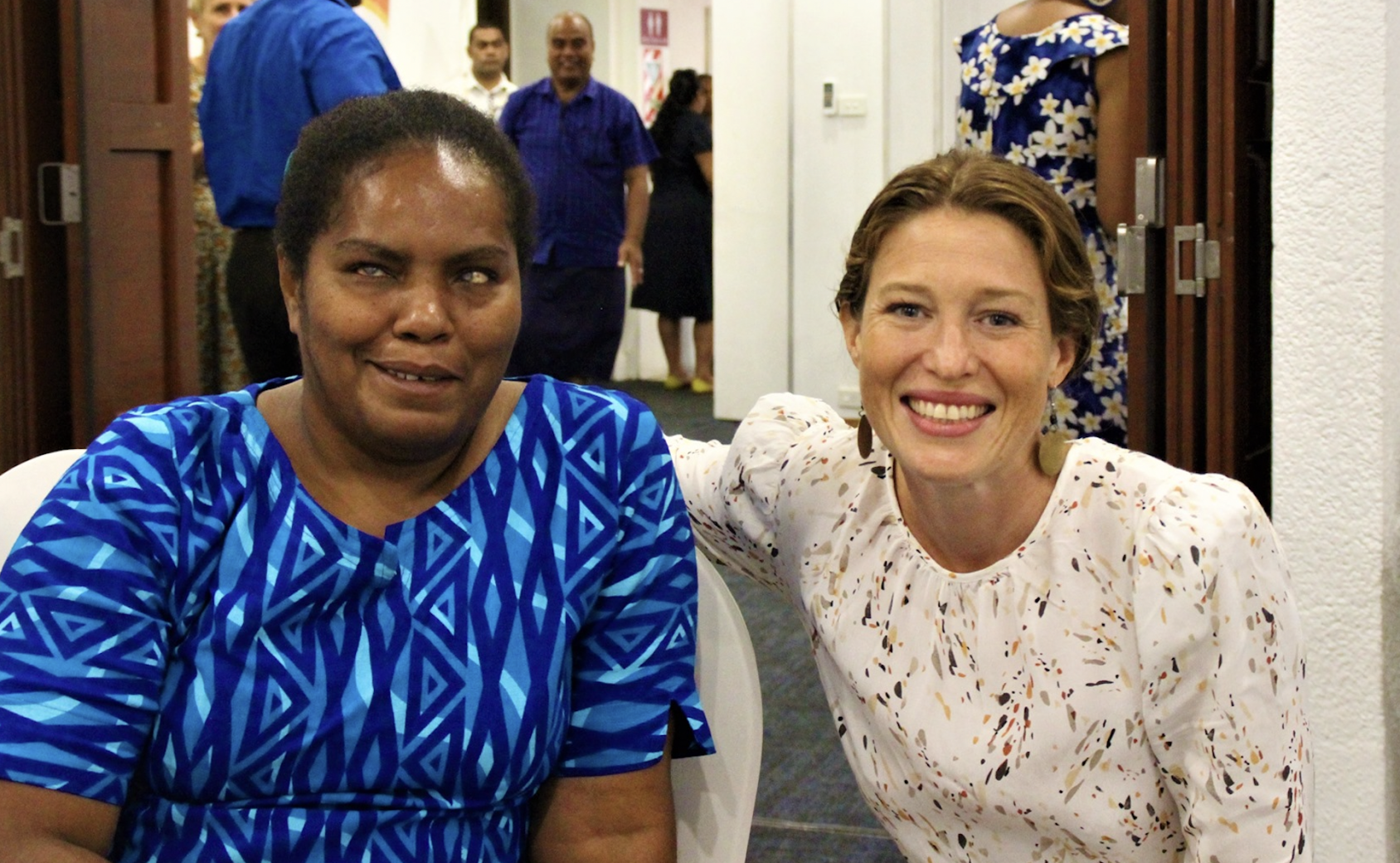 Two women smile directly at the camera one in a blue dress and one in a white dress. 