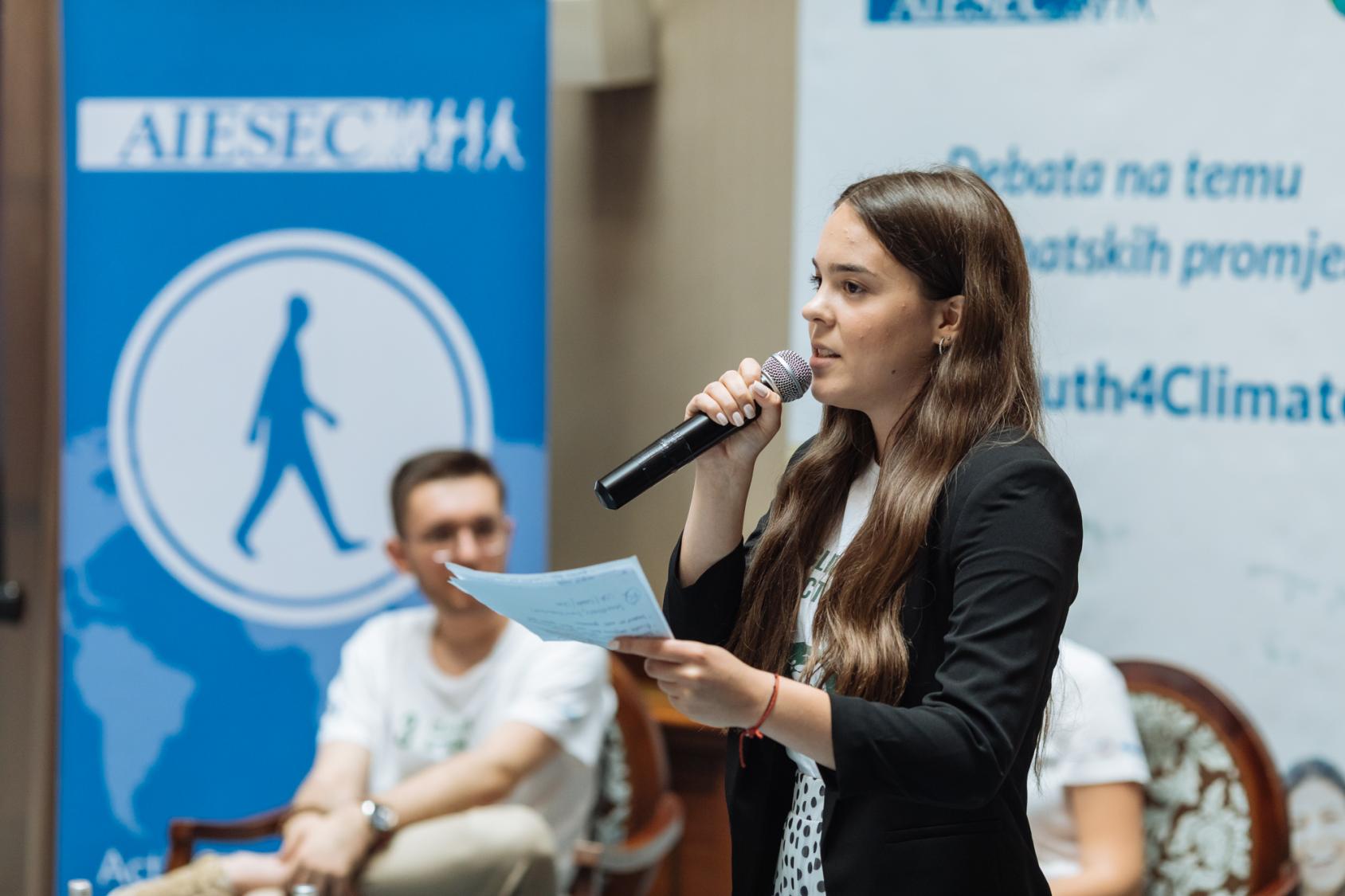 Une jeune fille en veste noire s'adresse à un public dans une salle avec un micro à la main.