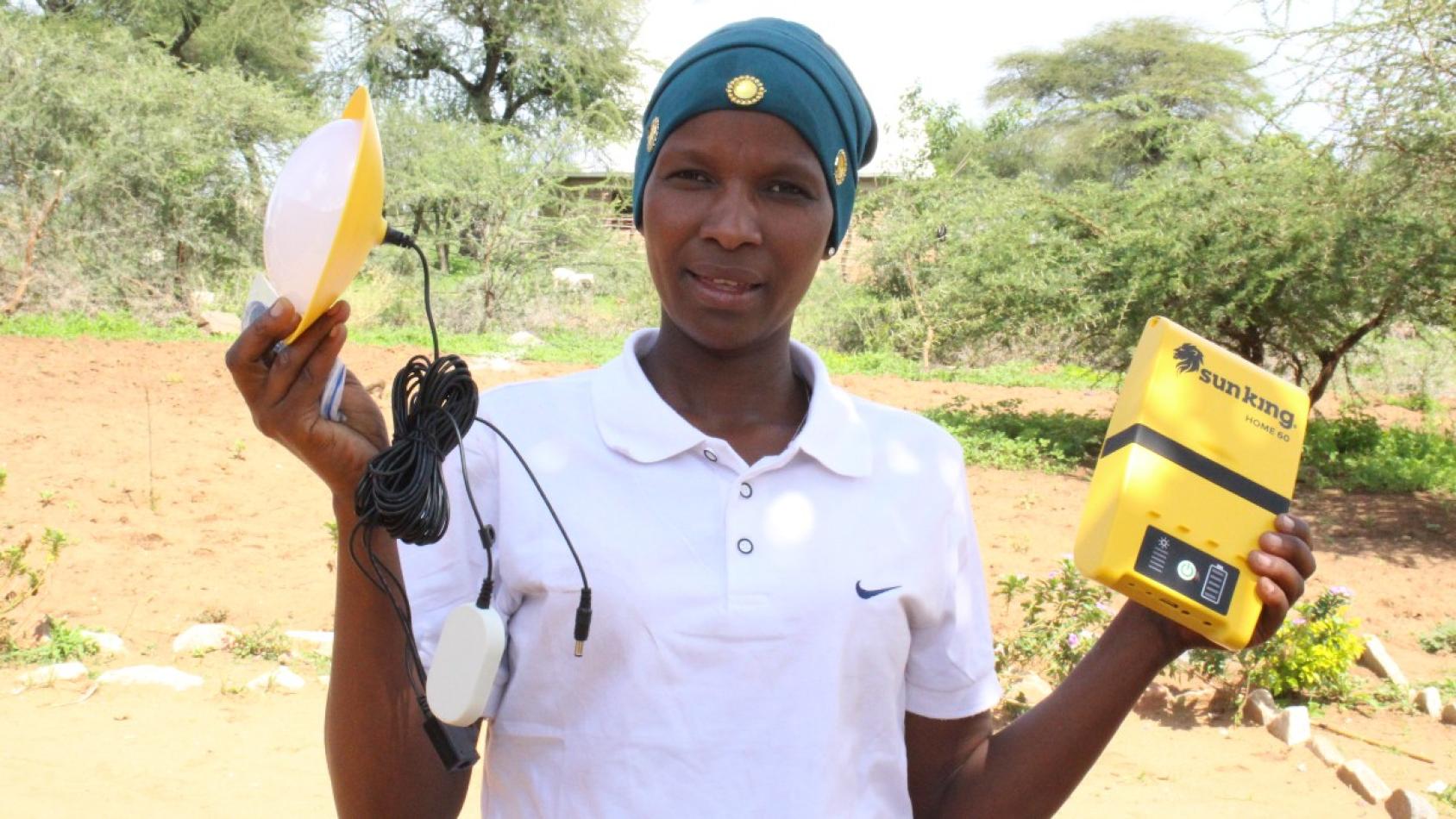 Une femme portant un polo blanc et un foulard serré autour de la tête se tient debout dans un champ, face caméra, une lampe dans la main droite et un équipement de production d’énergie solaire dans la main gauche.