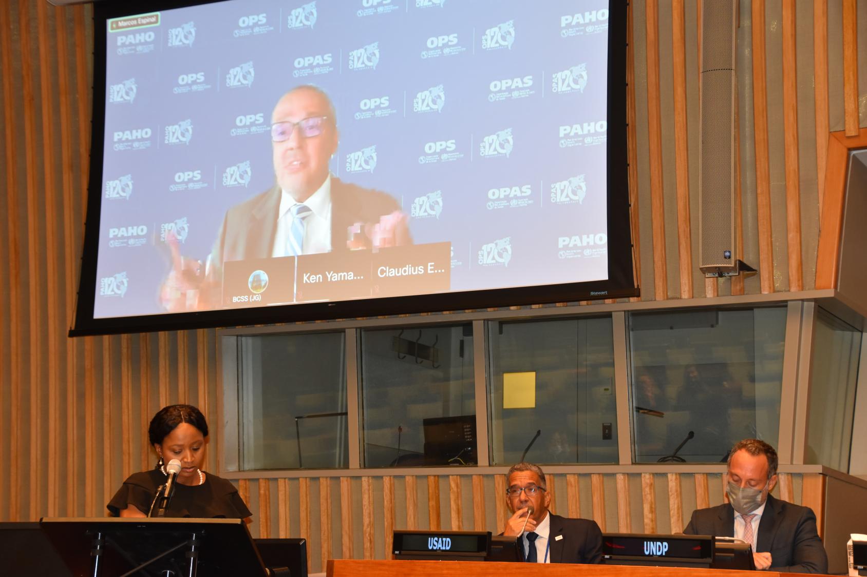 一名妇女对着话筒讲话，她身后的一个小组里有几个男性，一个大屏幕显示她身后也有一名官员在讲话。