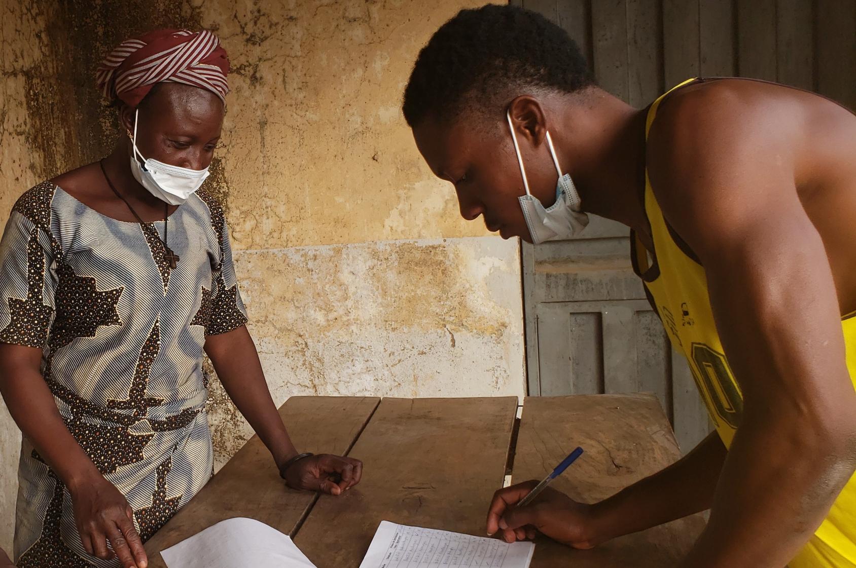 一个穿着黄色无袖衬衫的年轻男子正靠在一张木桌上，在一张纸上写字。一个女人站在桌子的另一边。