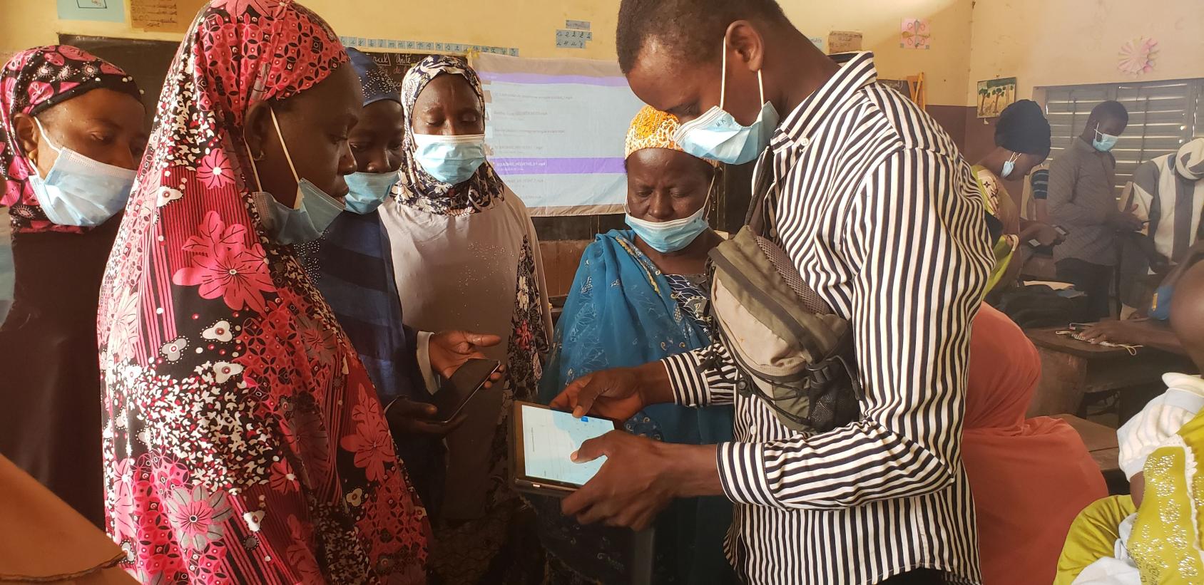 一个戴着面罩的年轻男子站在几个同样戴着面罩的妇女身边。他拿着一块平板电脑，向她们展示如何使用它。