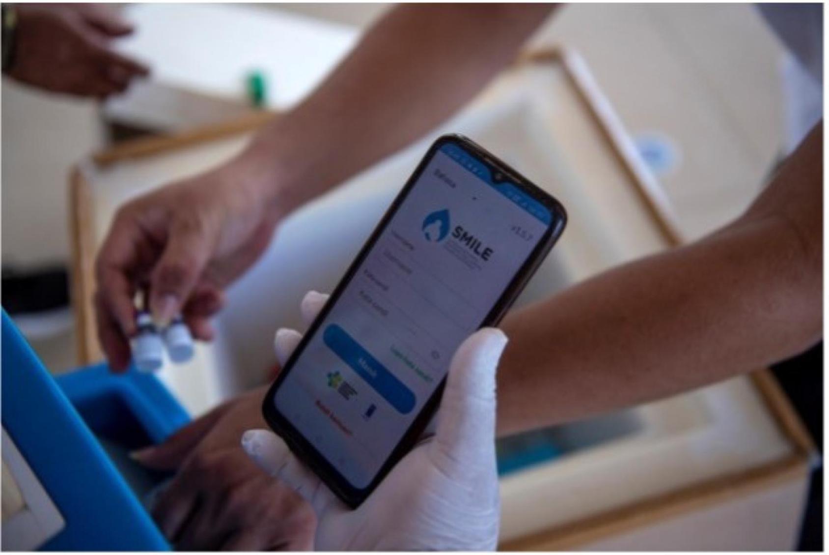 Gros plan sur un téléphone portable tenu par main gantée. Sur l'écran apparaît l'application SMILE, un dispositif de suivi électronique des données vaccinales et logistiques développé par le ministère indonésien de la Santé et le PNUD, avec l’appui du Gouvernement indonésien.