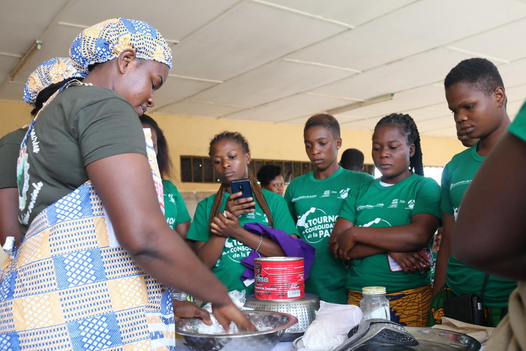 Une femme portant un tablier de cuisine se sert d'un ustensile devant des jeunes femmes qui l'observent faire avec attention.
