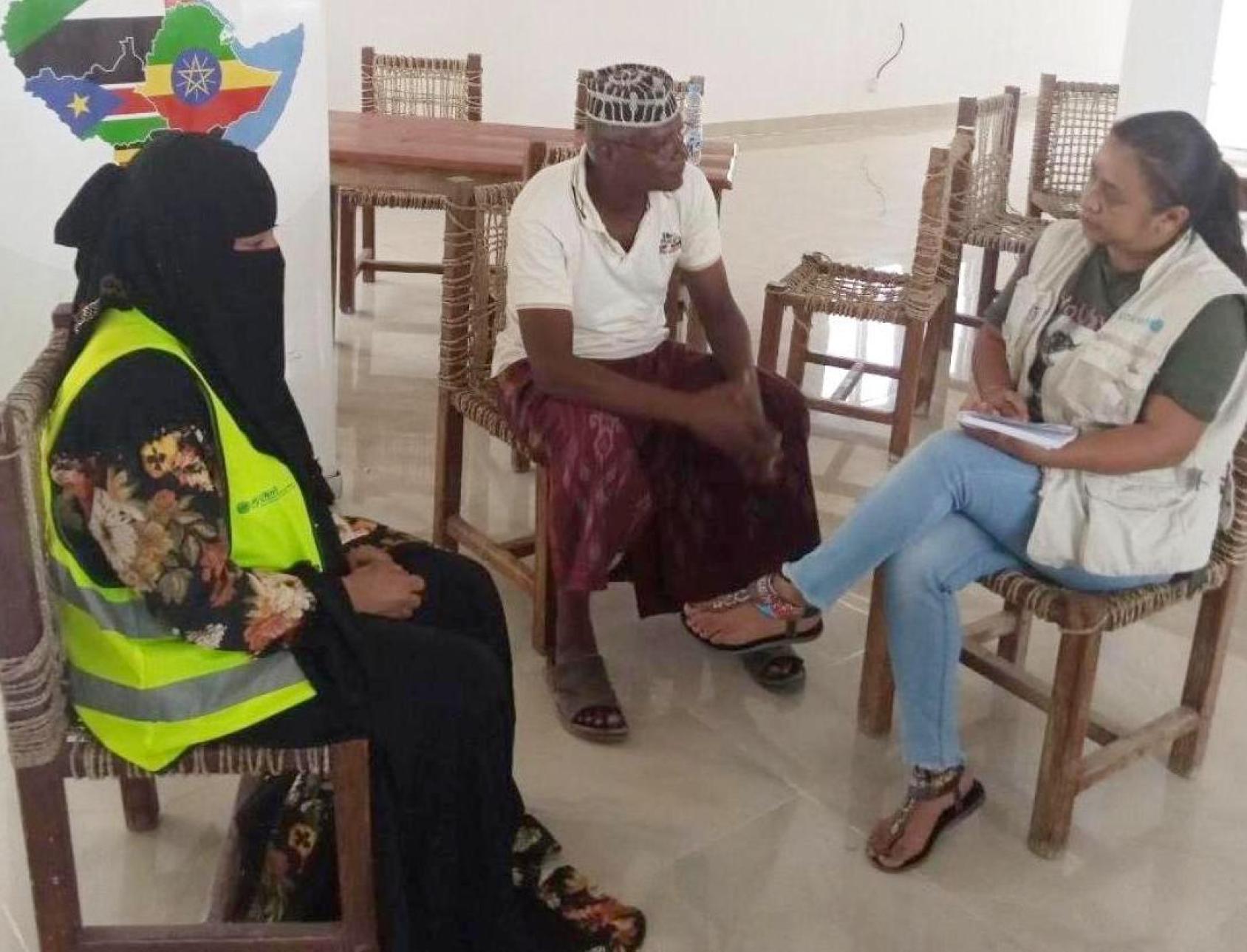 Un homme et une femme sont assis sur des chaises en bois dans une grande salle et discutent, tandis qu'une autre femme, qui porte un voile intégral noir, est assise avec eux et les écoute parler.