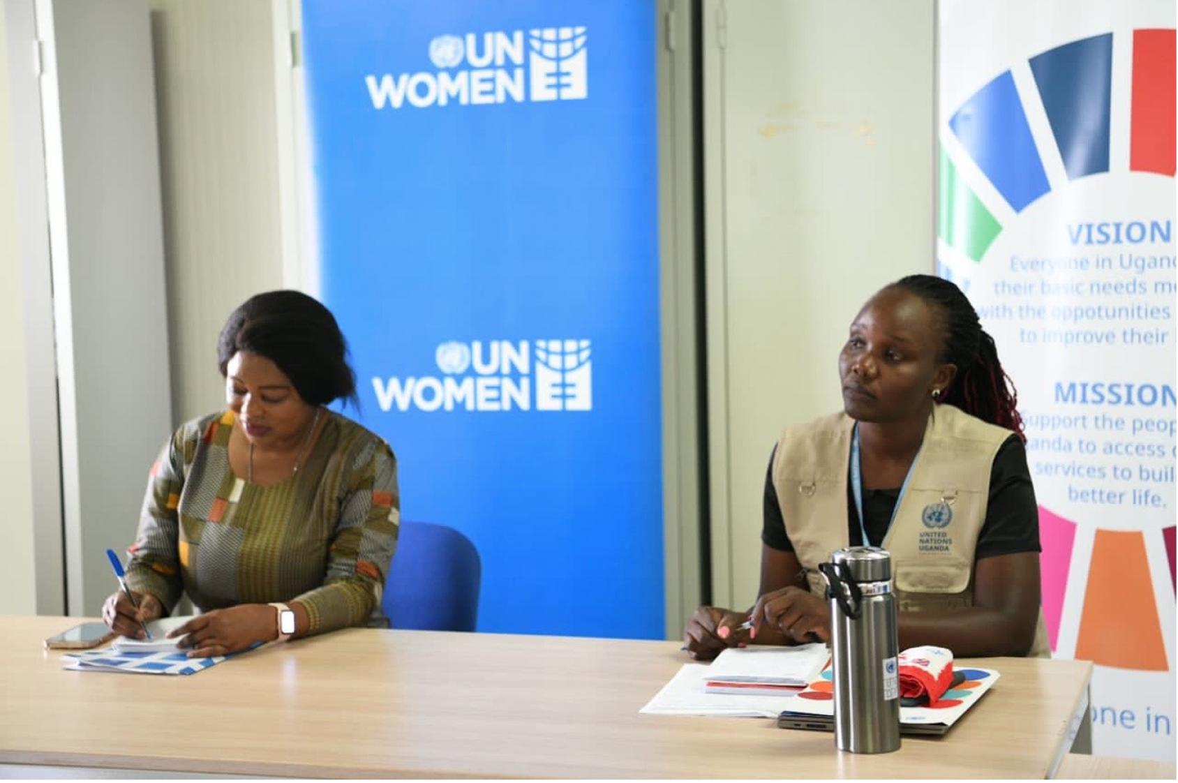 Two women, one wearing a UN Uganda vest, speak in front of a room, with UN Women and UNSDG banners behind them.