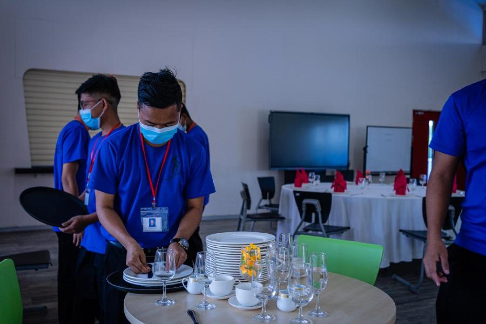 A group of teenaged-aged guys in blue t-shirts set up a venue, with glasses and table-cloth lined tables.