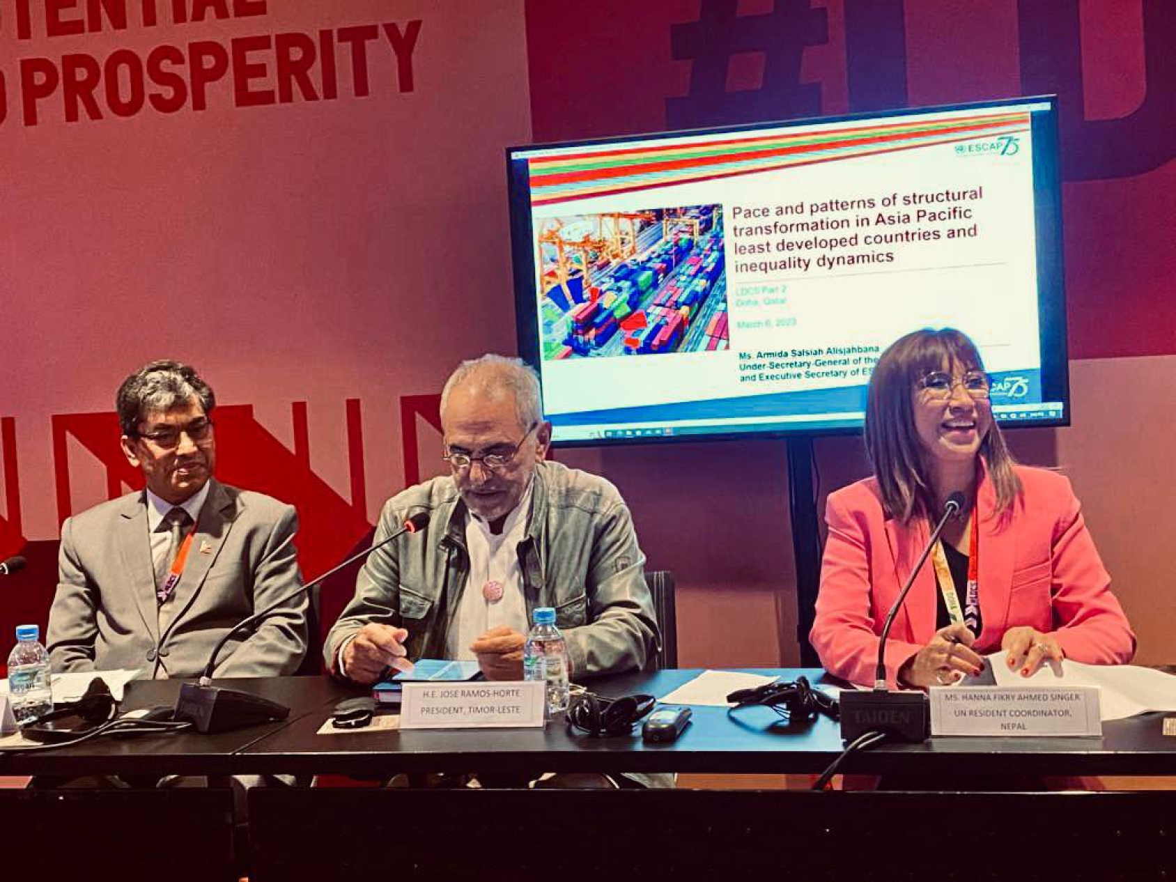 UN officials at a conference speak into their mics, with bright signs behind them.