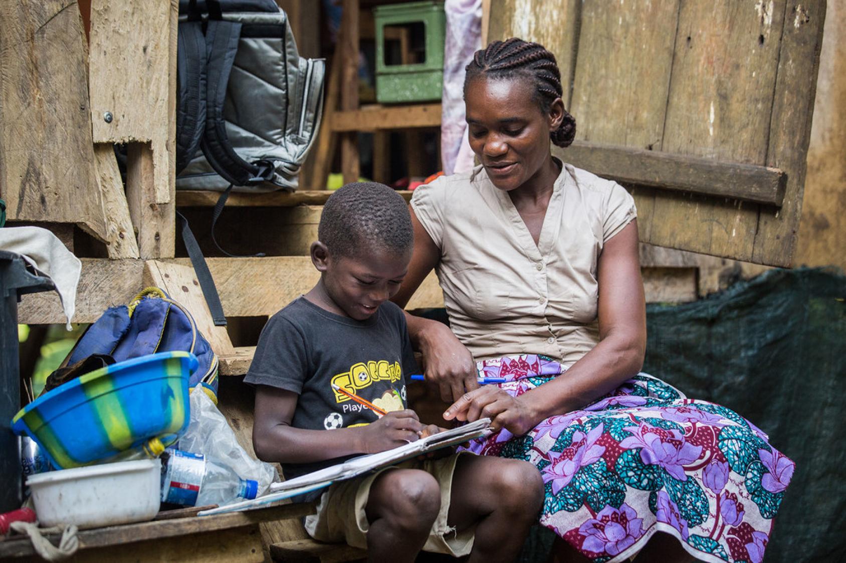 A woman in a beig top and flowered skirt helps a young boy with school work. They are sitting outside in front of a structure.