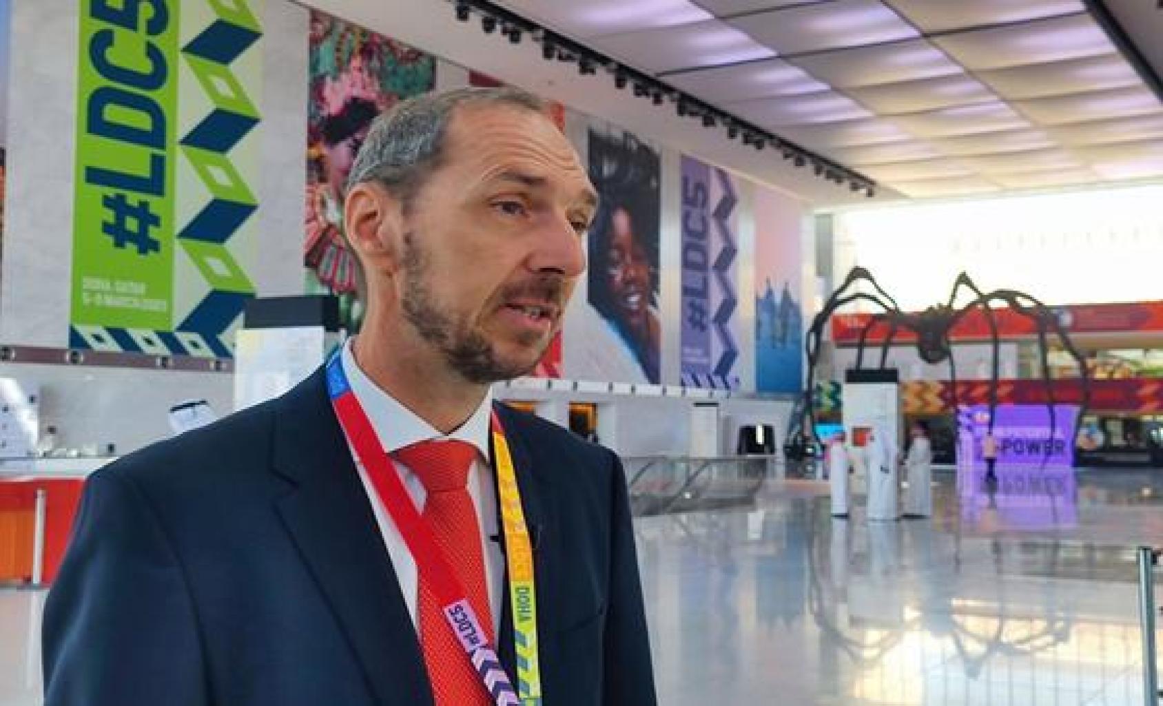 A man in a dark suit and white shirt stands before LDC posters in a hall.