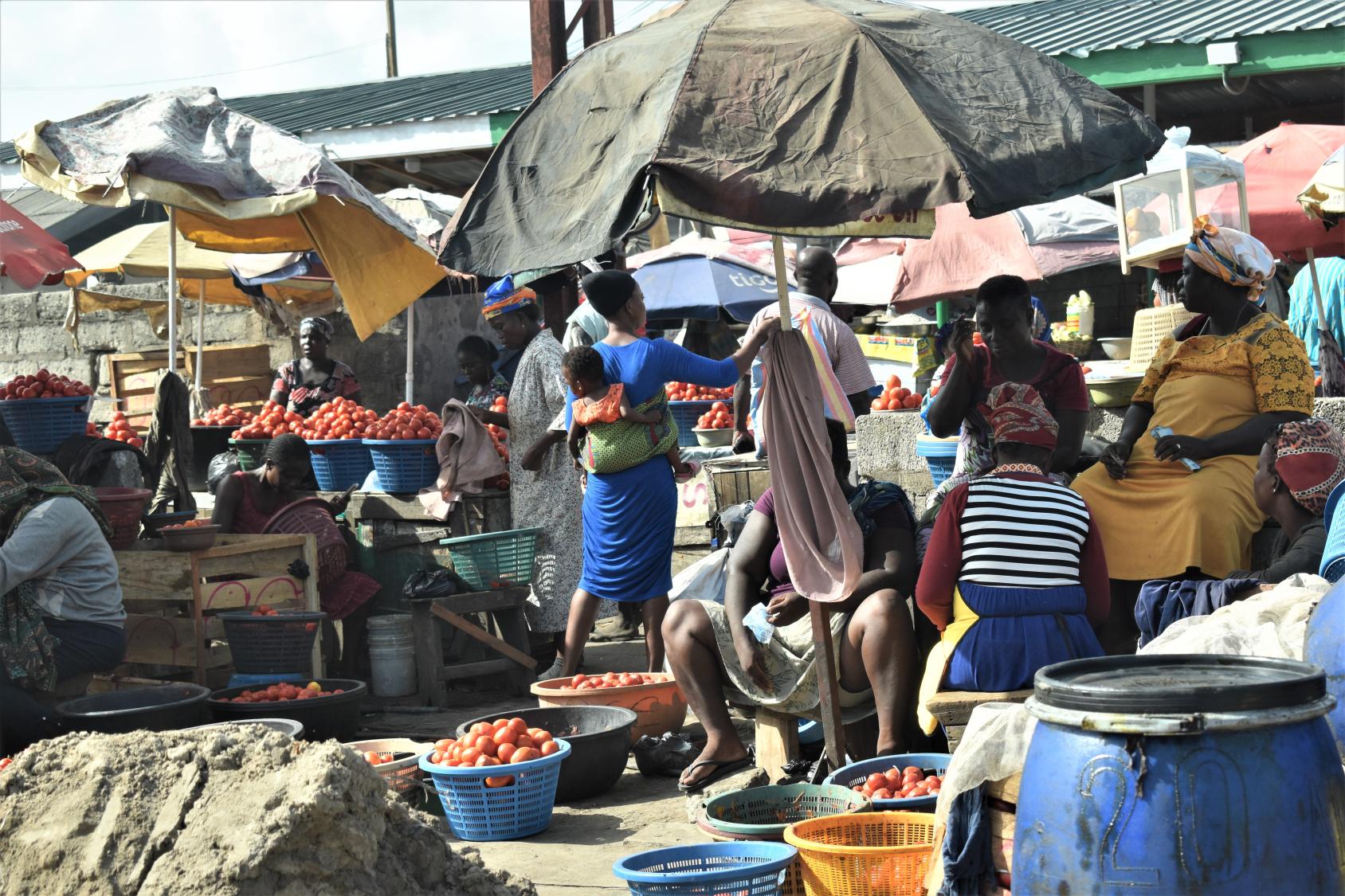 A market scene with men and women standing and surrounded by baskets with vegetables and fruits under umbrellas.