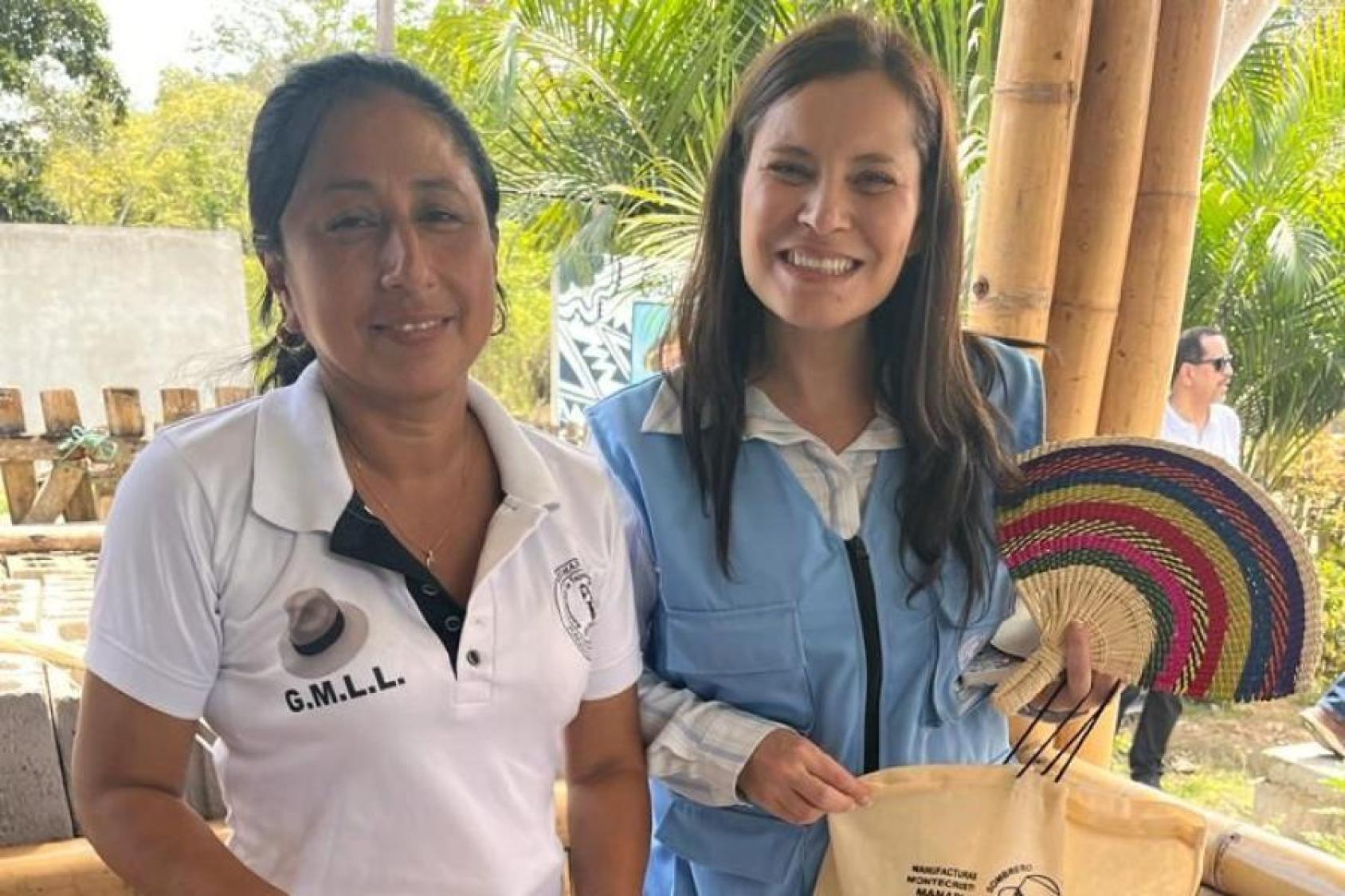 Two women, one in a blue shirt on the right and the one on the left in a white t-shirt; the one in blue is carrying a traditional straw fan and a cloth bag