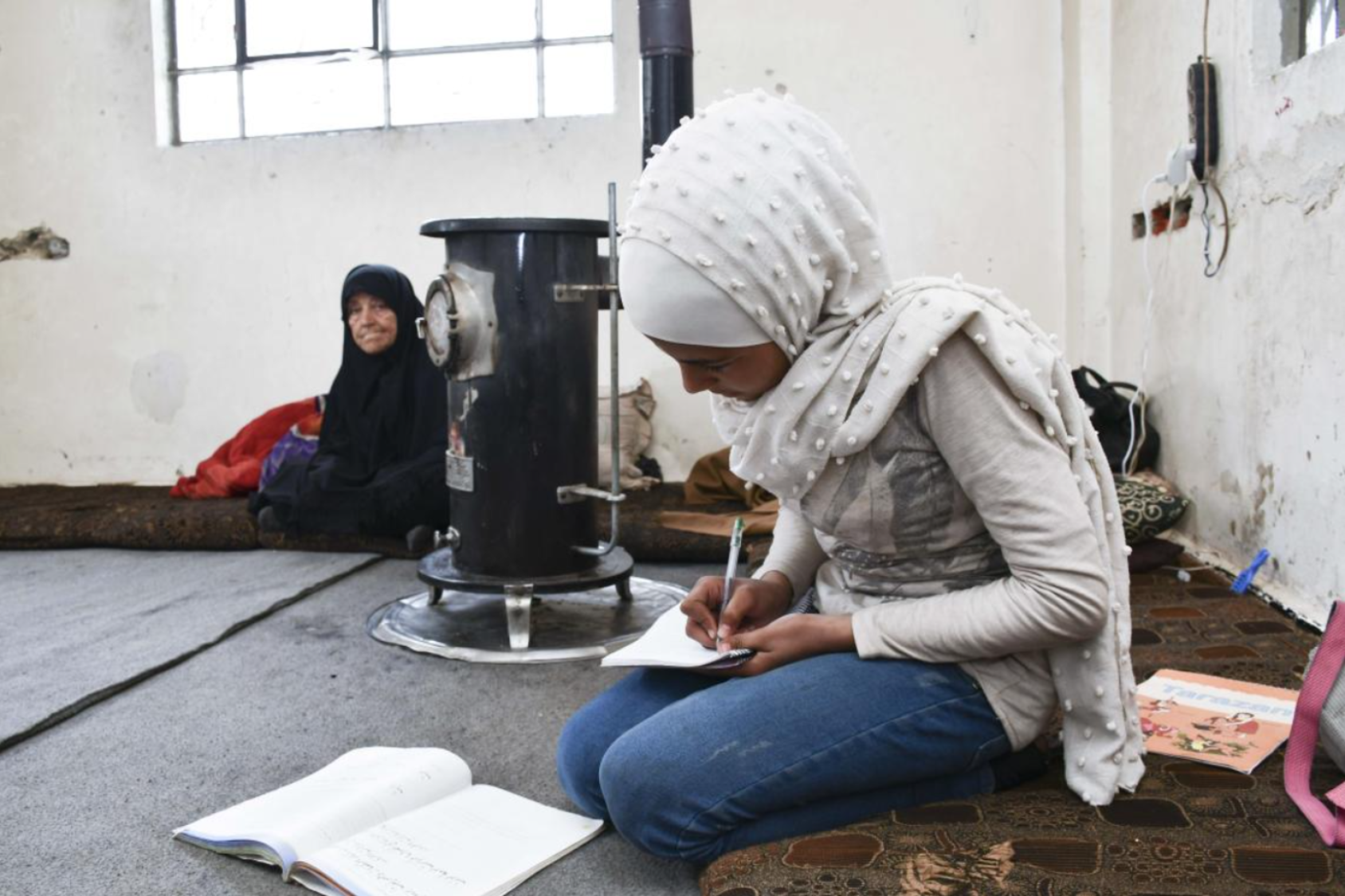 A young girl in a white dress and headscarf crouches on the floor where she writes in a notebook while another girl watches her in the background