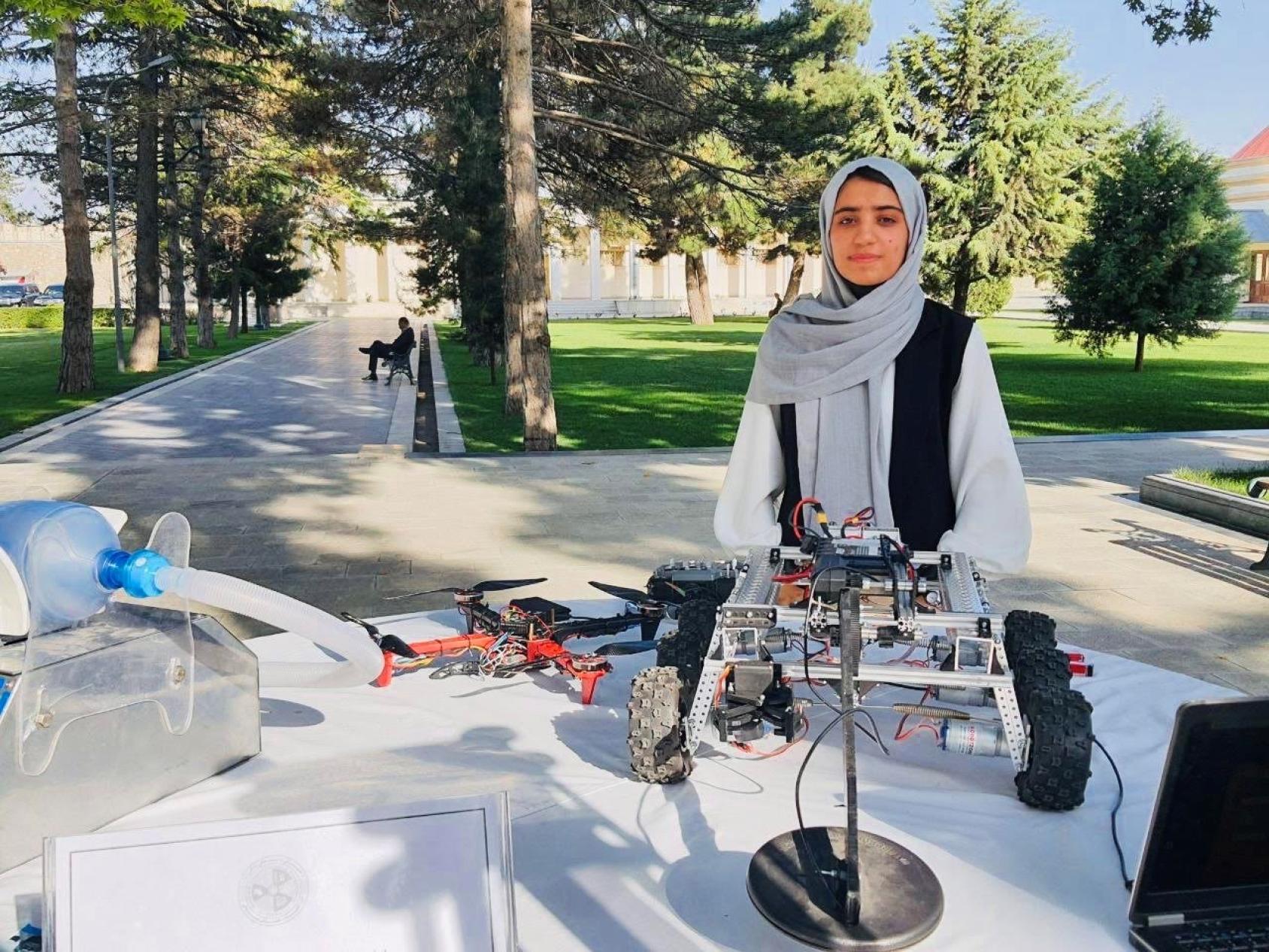 An Afghan girl in a white and black dress and headscarf stands behind a table on which a model of a vehicle with robotic parts is displayed