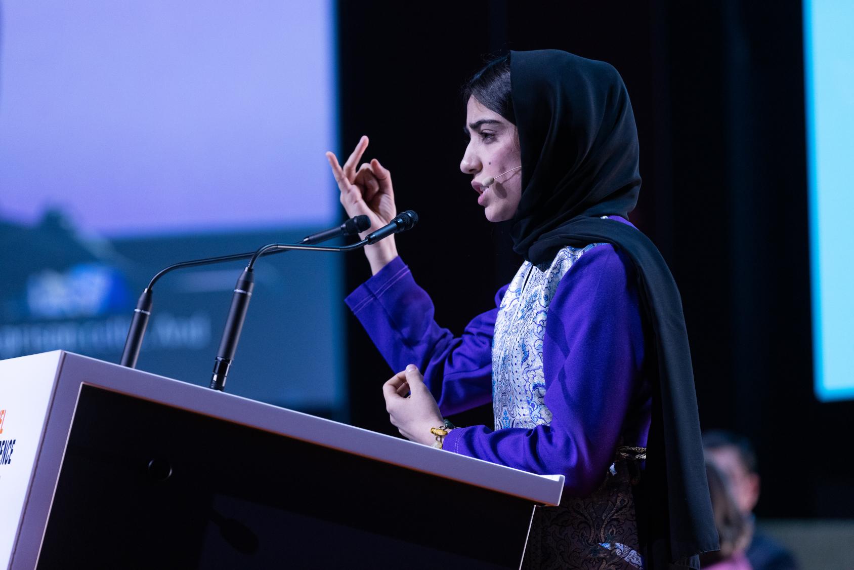 A girl in a purple dress and headscarf speaks on a podium