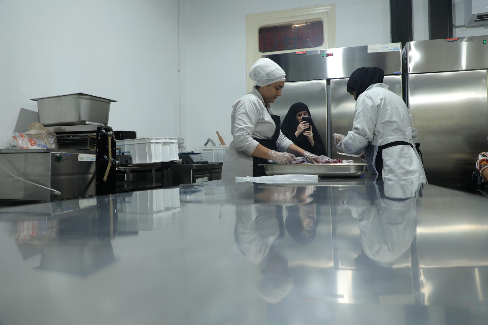 Women in chef's uniforms in a large kitchen slice up meat. 