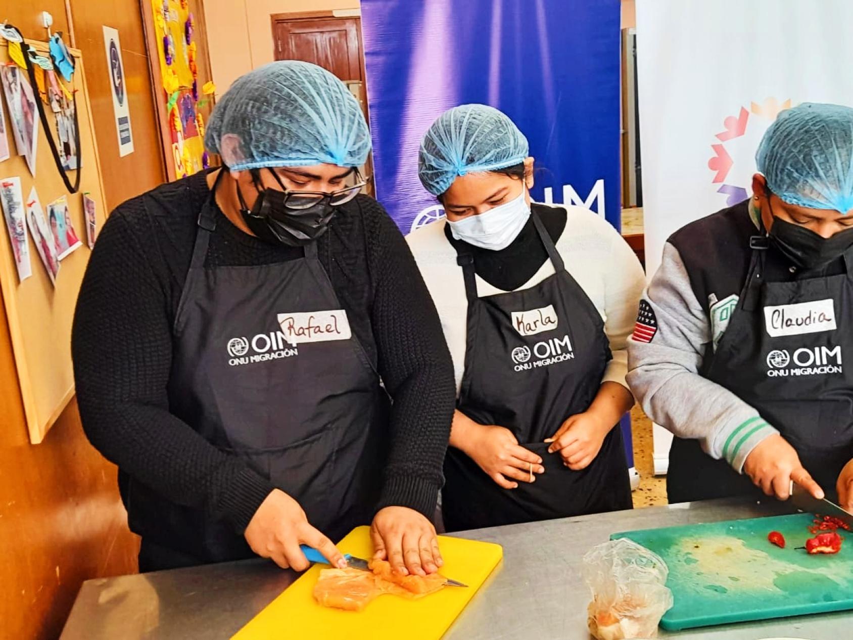 Three chefs in black aprons stand over a table as one of them cuts through fish