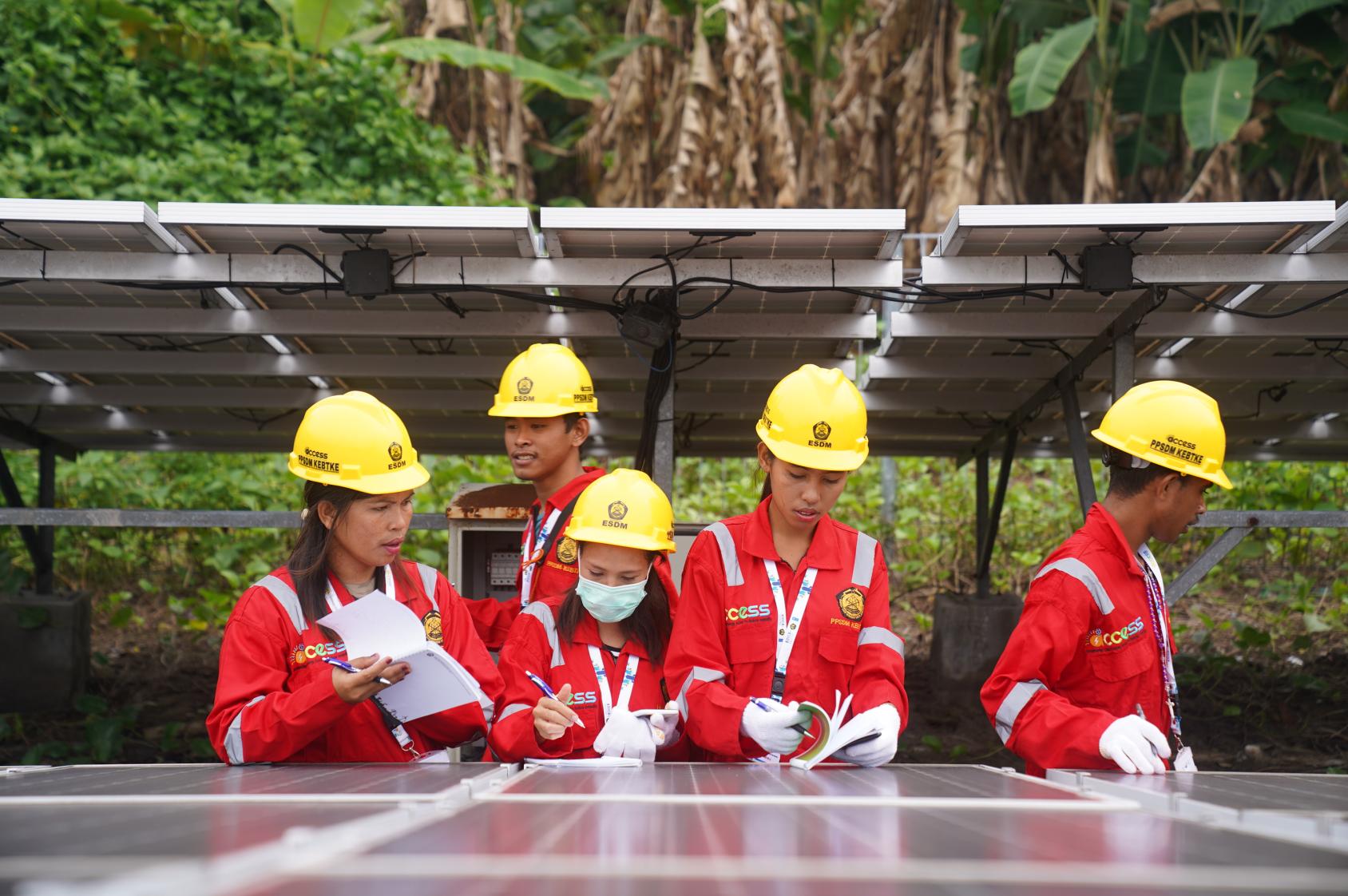 A group of five workers in red suits and yellow hardhats stand over a solar panel