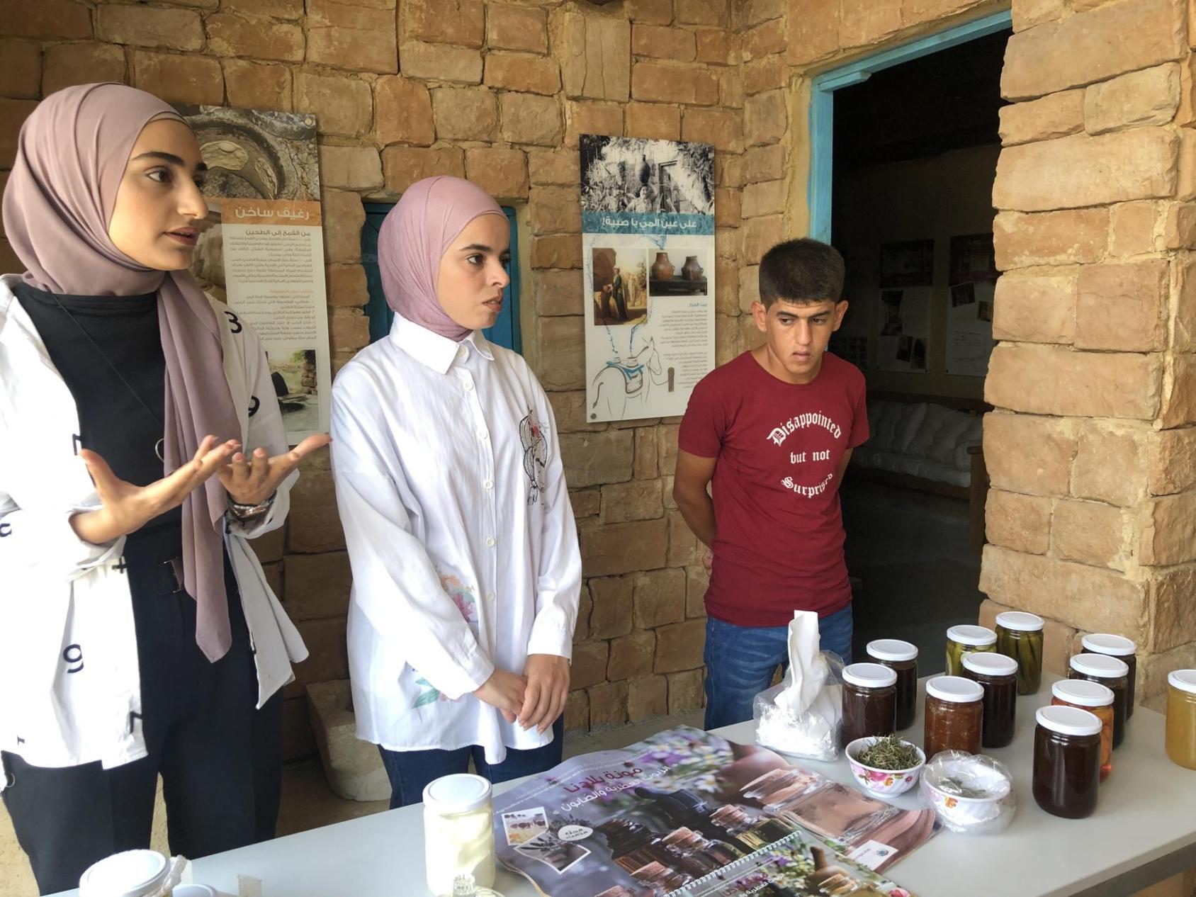 Three young people, two girls in a white dress and a pink headscarf and a boy in a red shirt stand in front of a table that carries small bottles and jars of various products