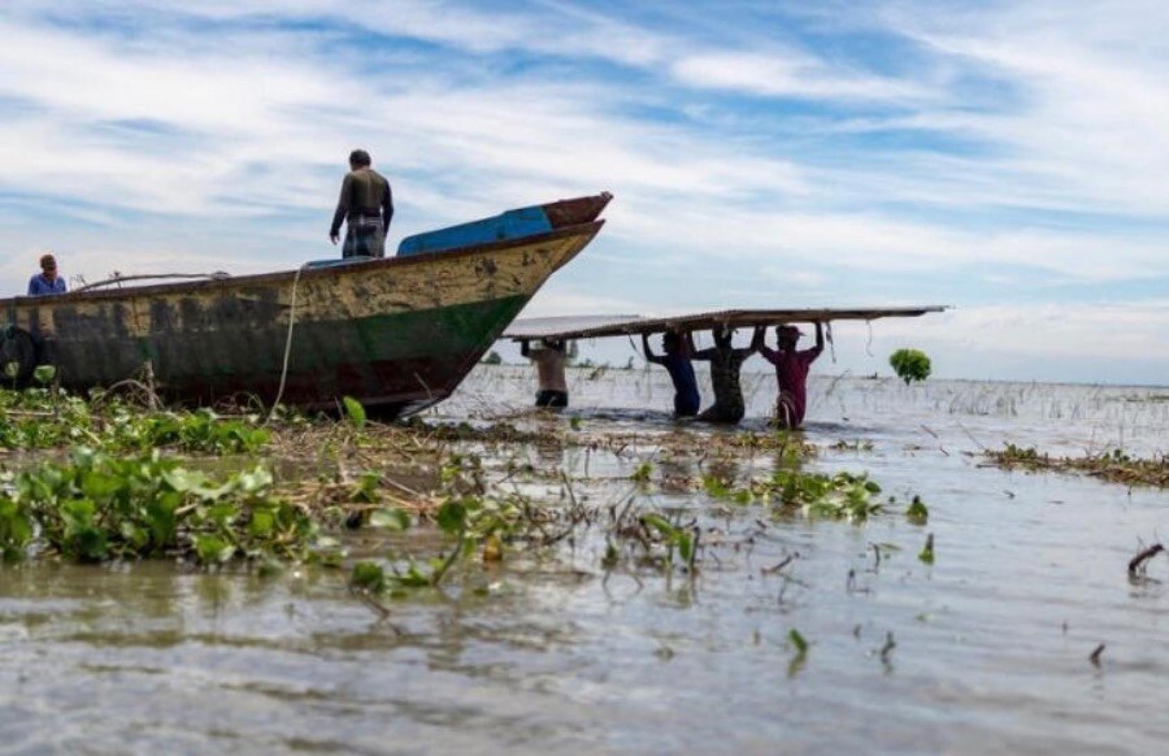 A silhouetted person stands atop a boat while more carry a board near a river bank.