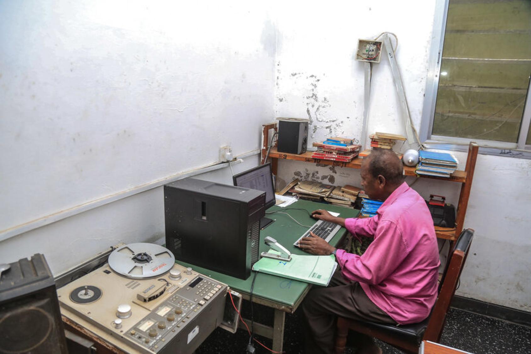 A man in a pink shirt sits at a computer desk typing on a keyboard. Next to him is a table with a grey coloured audio recorder