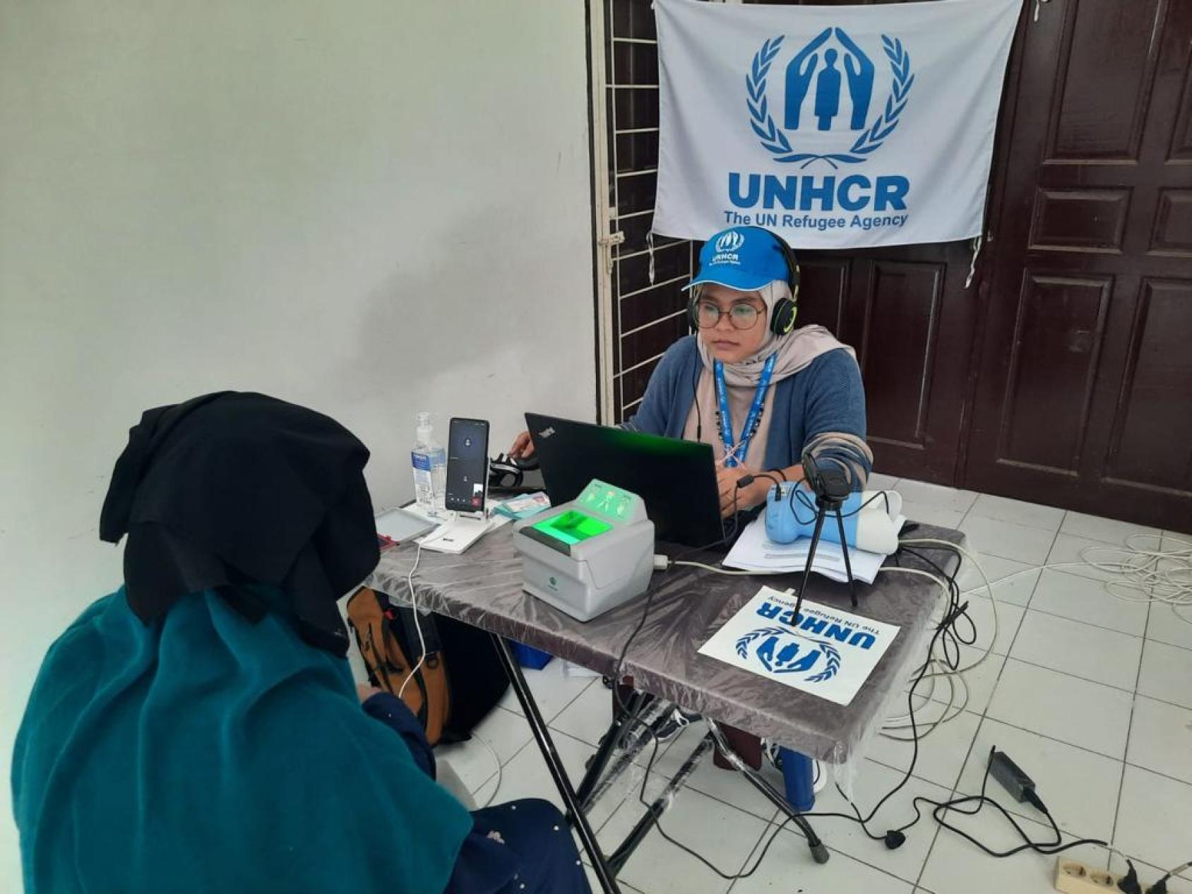 A woman in a blue cap sits at a desk with a computer interviewing a woman dressed in blue and black.