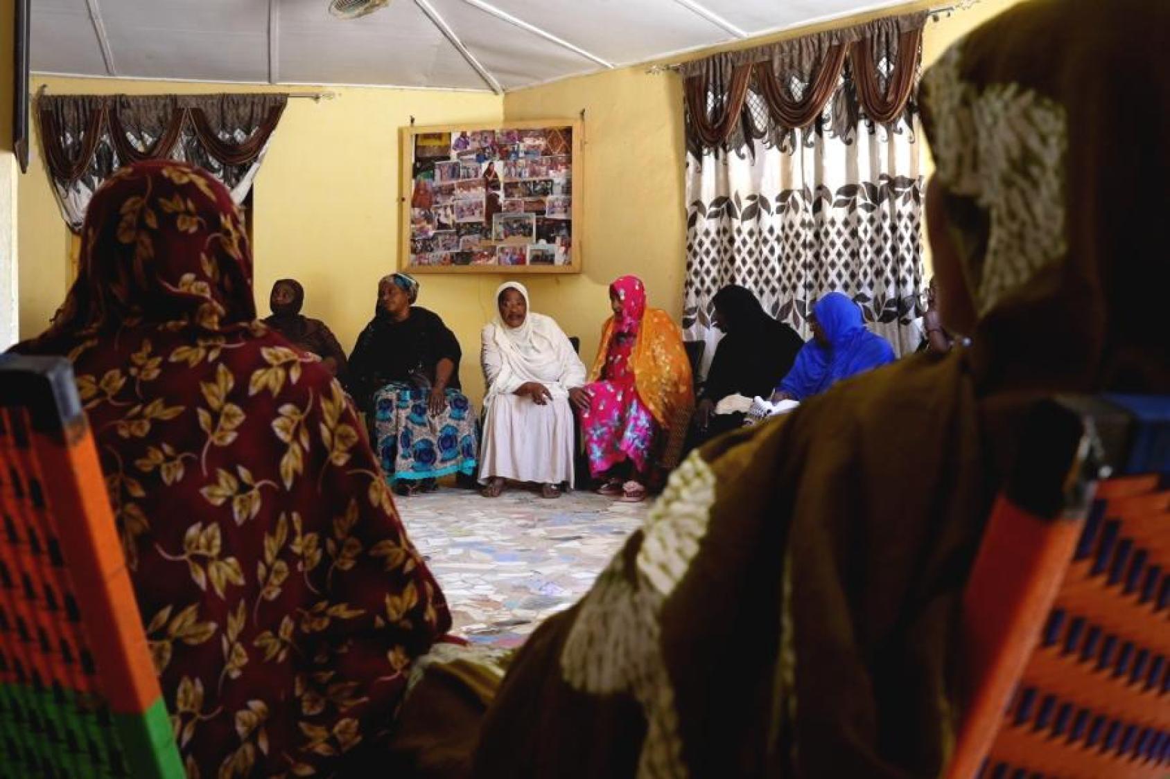 A group of women in colourful clothes sit in a room with yellow walls, discussing and talking.