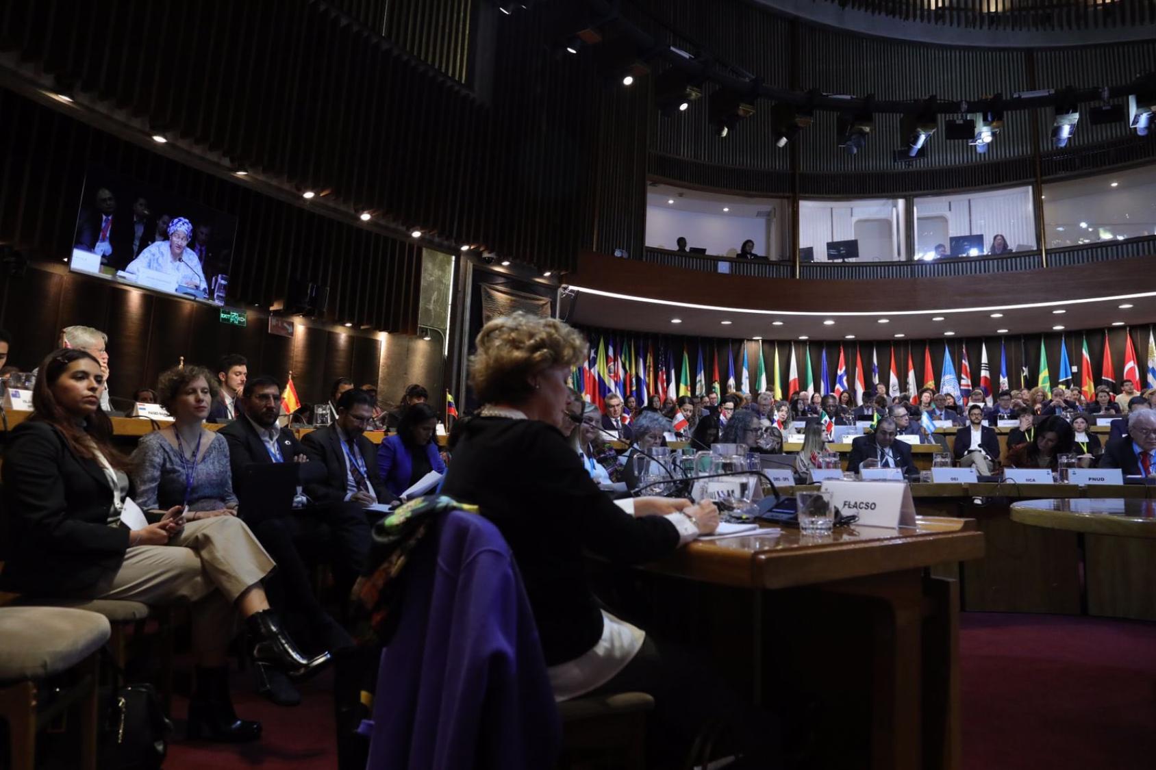 A group of people sitting in a darkened meeting room across a podium, seemingly in discussions.