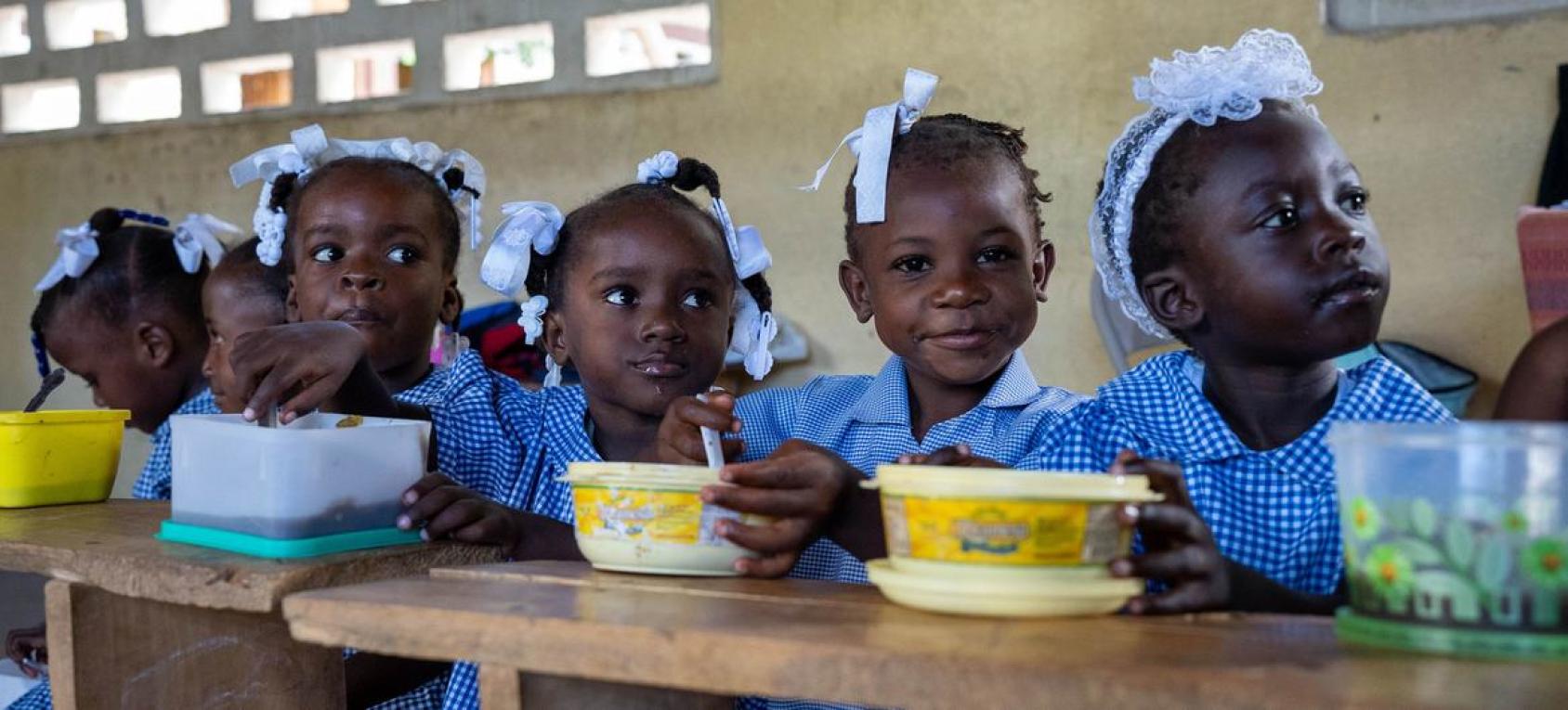 Five children in blue school uniforms and white ribbons sit on a desk with their lunch boxes open