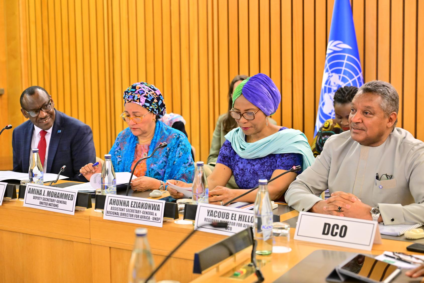 A row of people sitting at a podium with microphones. From left to right, a man in a black suit, followed by two women in colourful dresses and headscarves, followed by another man in a grey shirt. Behind them is a wooden conference wall with a blue UN flag.