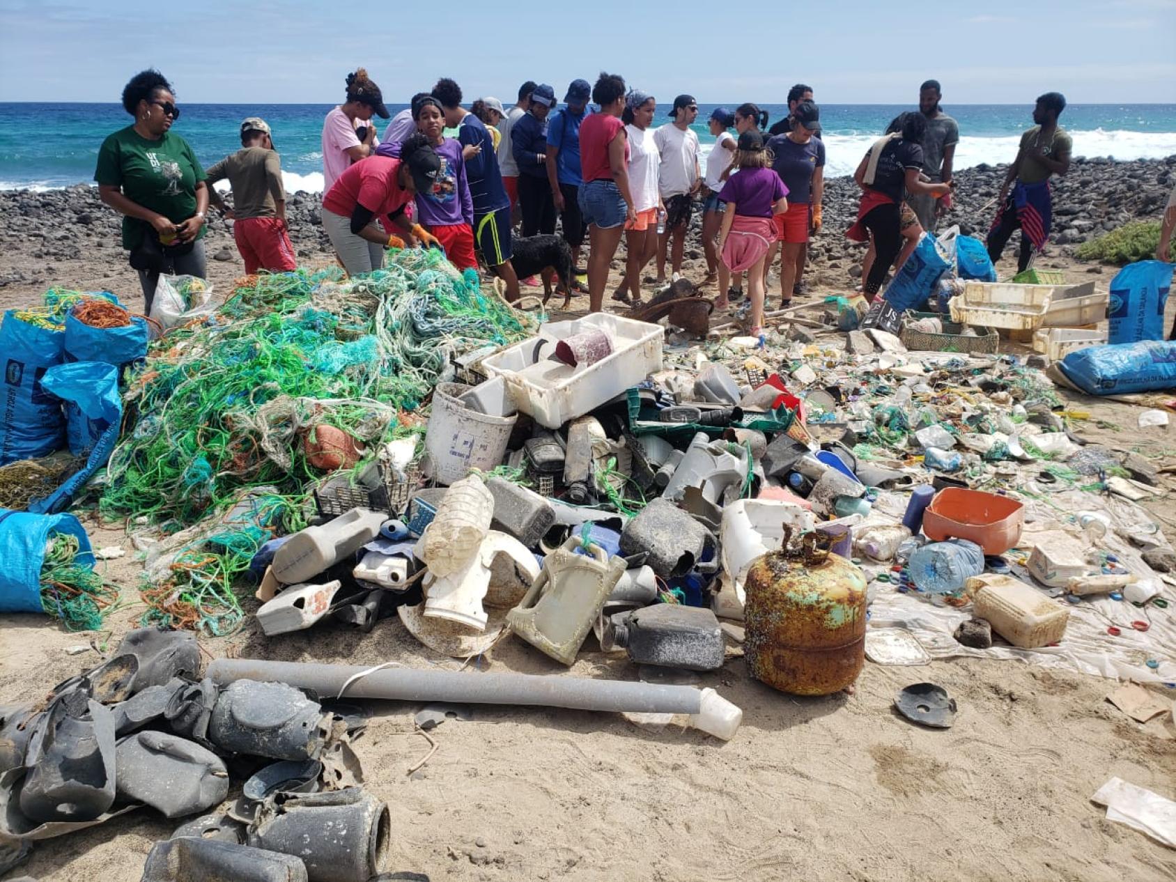 A group of people gather on a beach where numerous plastic waste products are lying strewn at their feet.