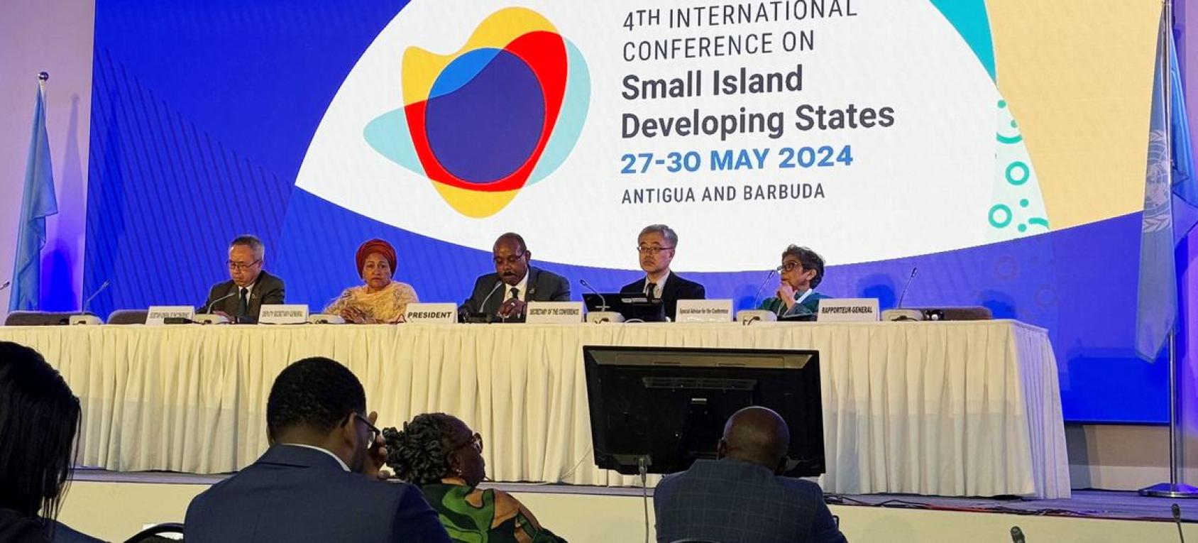 A conference setting with a panel of five individuals seated at a long table on a stage, facing an audience. The backdrop displays the text “4TH INTERNATIONAL CONFERENCE ON SMALL ISLAND DEVELOPING STATES 27-30 MAY 2024 ANTIGUA AND BARBUDA” along with a colorful logo