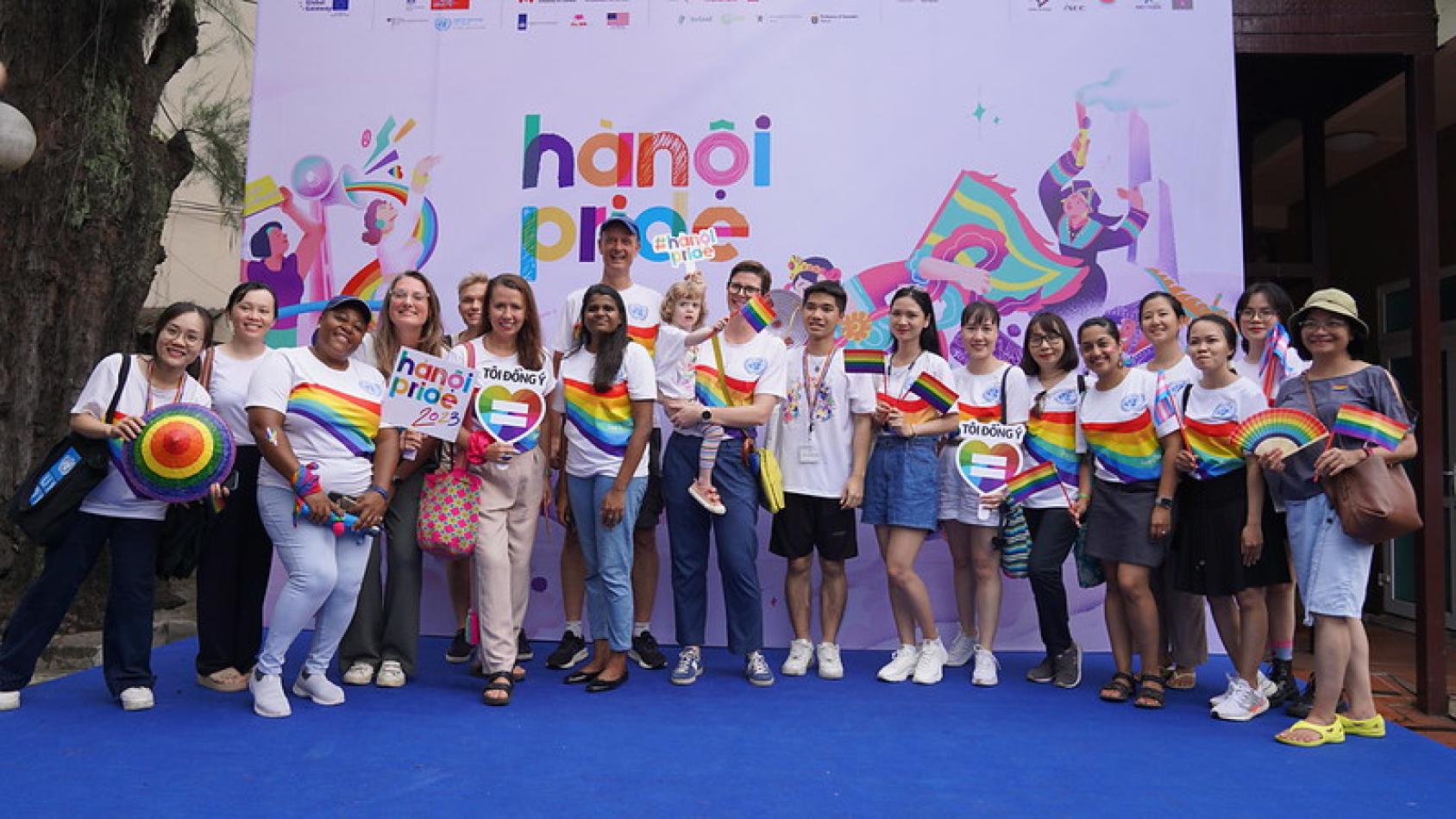 A group of people wearing colourful pride t-shirts in front a sign that reads 'Hanoi pride'