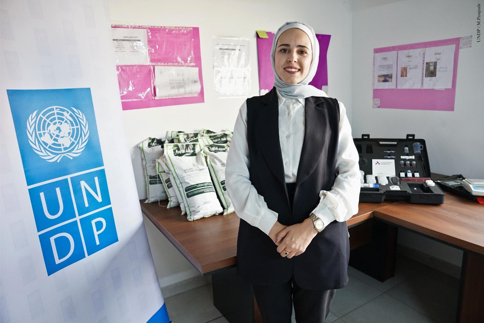 A woman in a black dress and white headscarf smiles to the camera