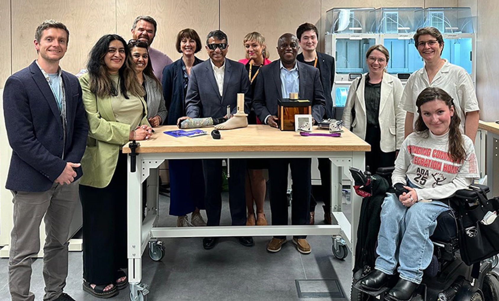 A group of people in suits and some wearing dark glasses and in a wheelchair stand around a desk with various devices in front of them.