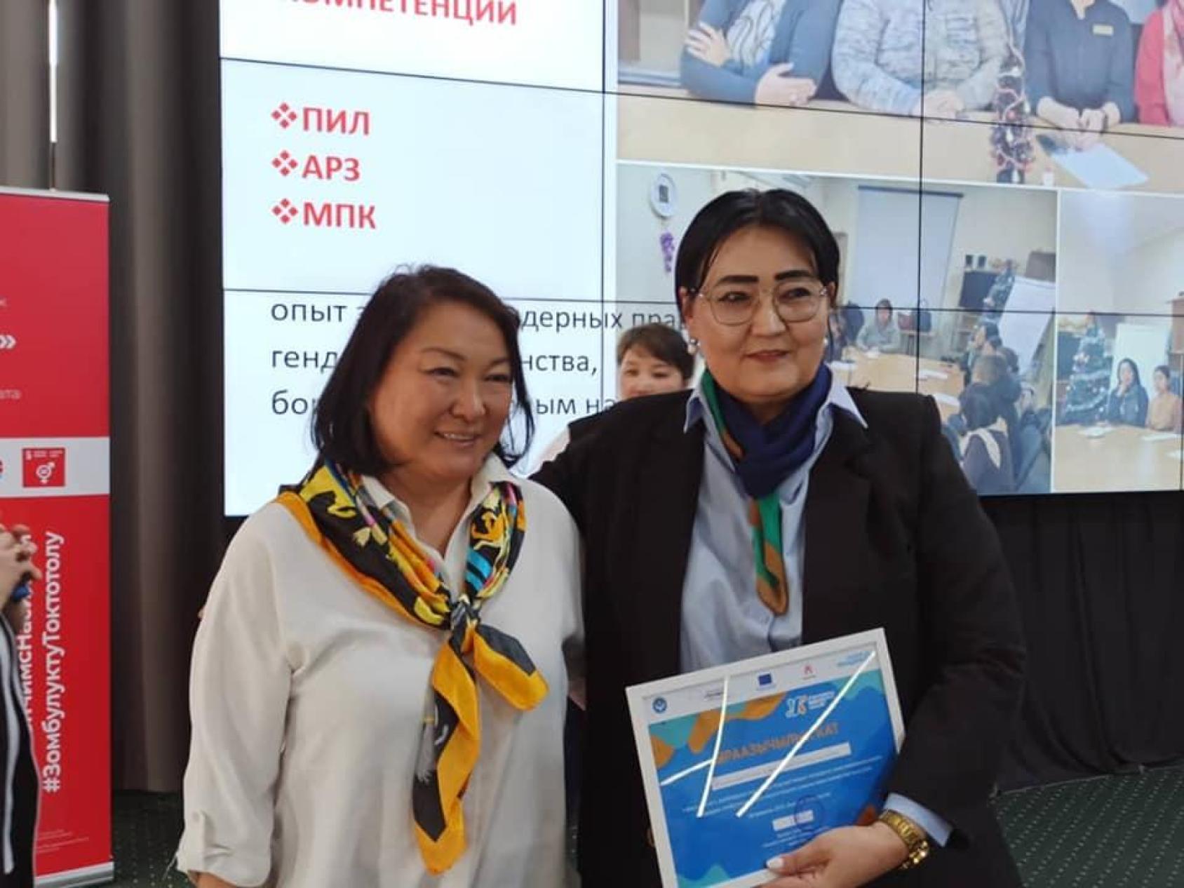 Two women stand next to each other, one in a white shirt and yellow scarf and the other in a navy blue blazer holding a folder
