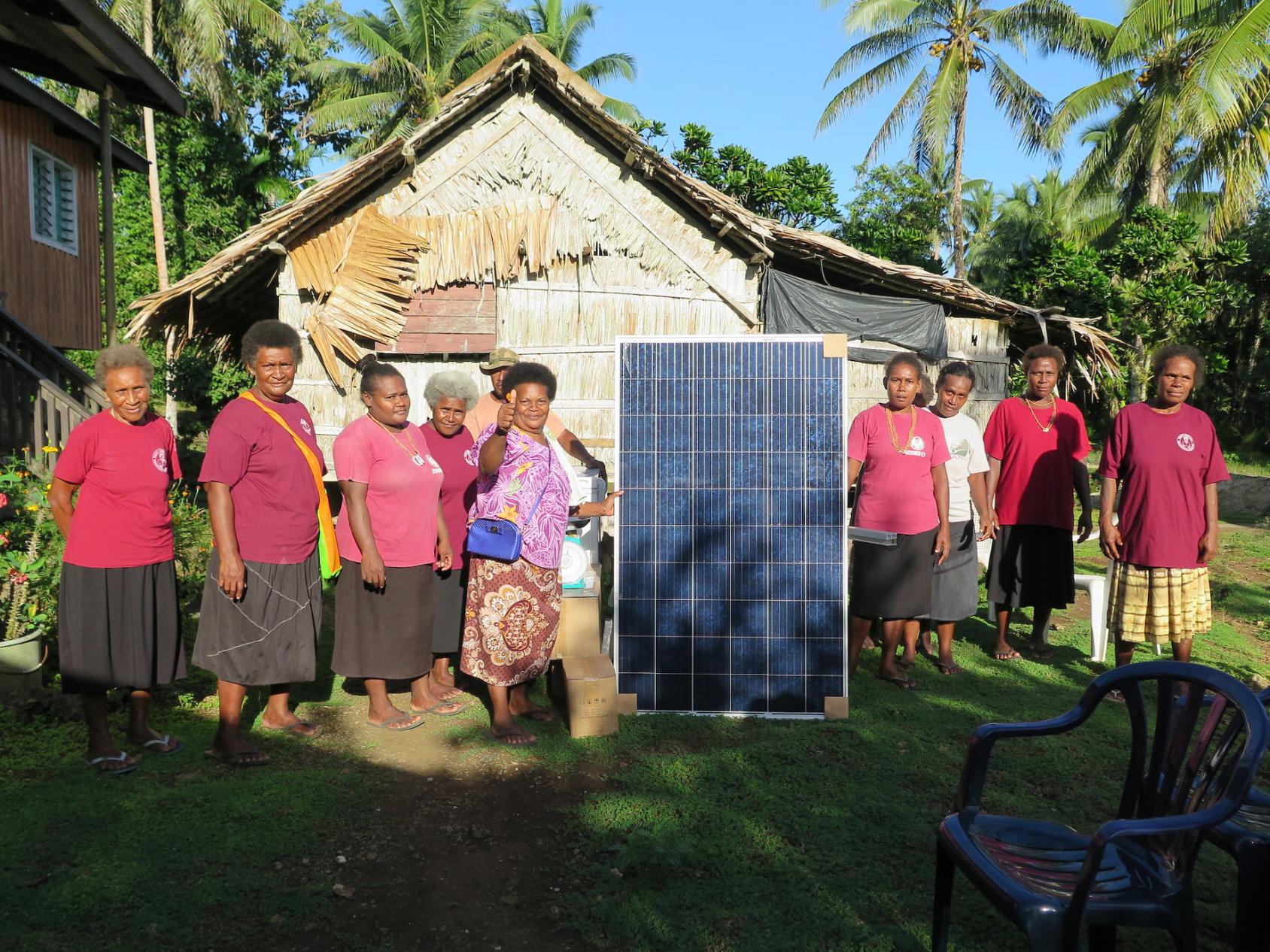 A group of women in red shirts stand in front of a big solar panel of a house in the Solomon Islands