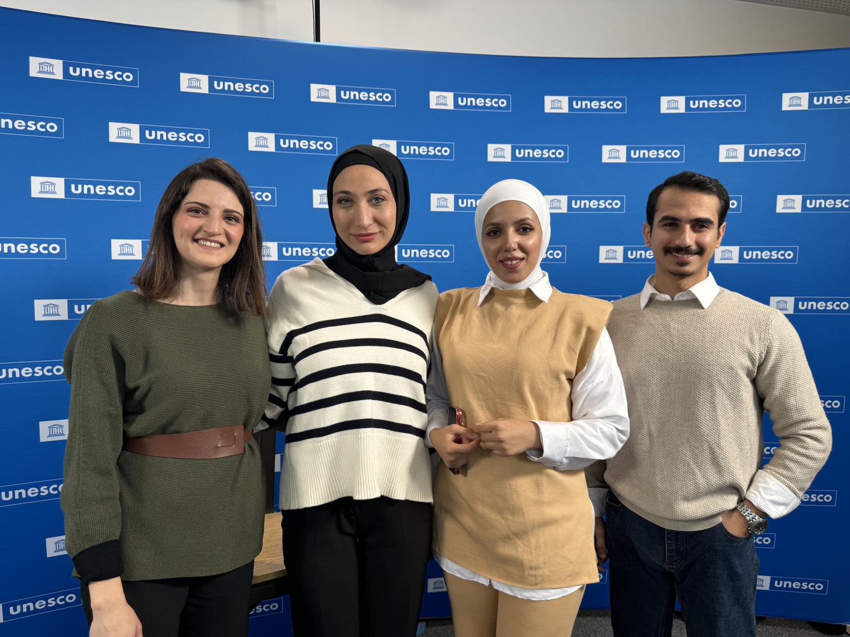 A group of three women and one man stand in front of a backdrop that has the UNESCO logo on it 