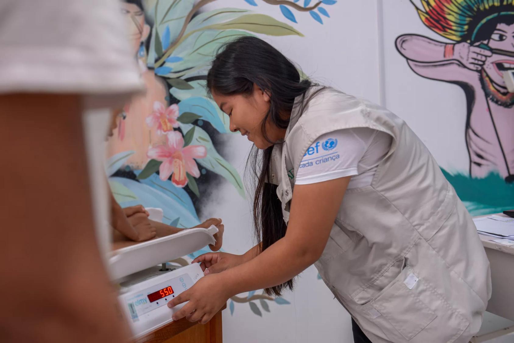 A woman in a grey vest looks over a child on a weighing scale