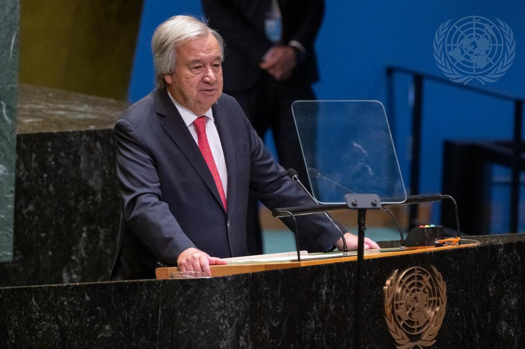 UN Secretary-General Antonio Guterres in a black suit and red tie stands at a UN podium for a speech