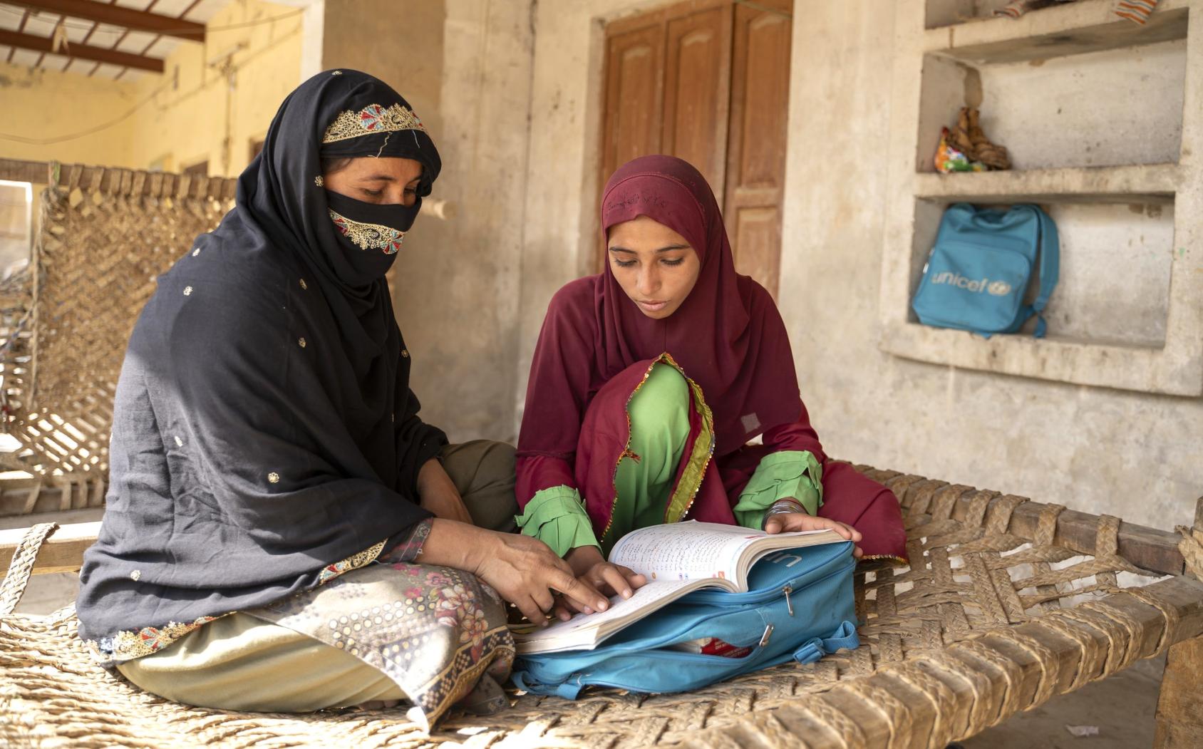 An older woman in a black headscarf with her face covered sits with a young girl in a red dress reading a book that is open on their laps.