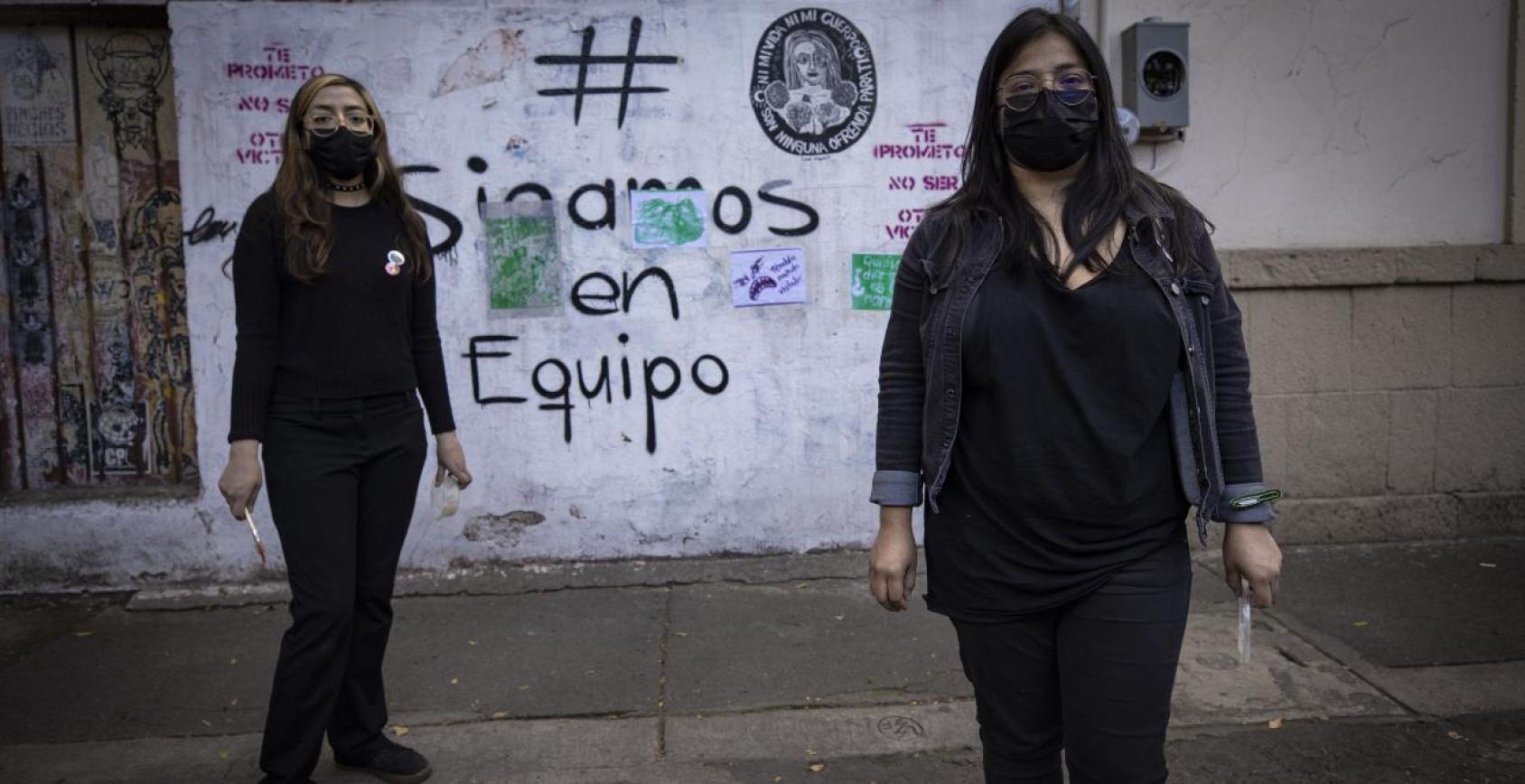 Two women in black clothes and masks stand in front of a graffiti wall drawing attention to sexual violence