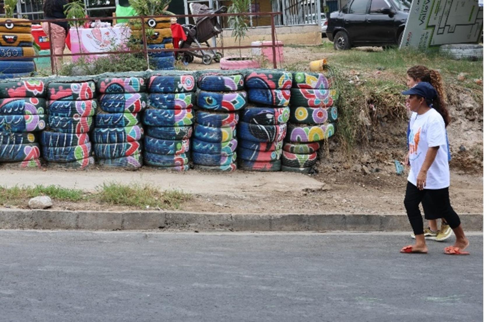 Two women walk in white tshirts and black pants walk down an asphalt road. In the background are rows of colourful tires.